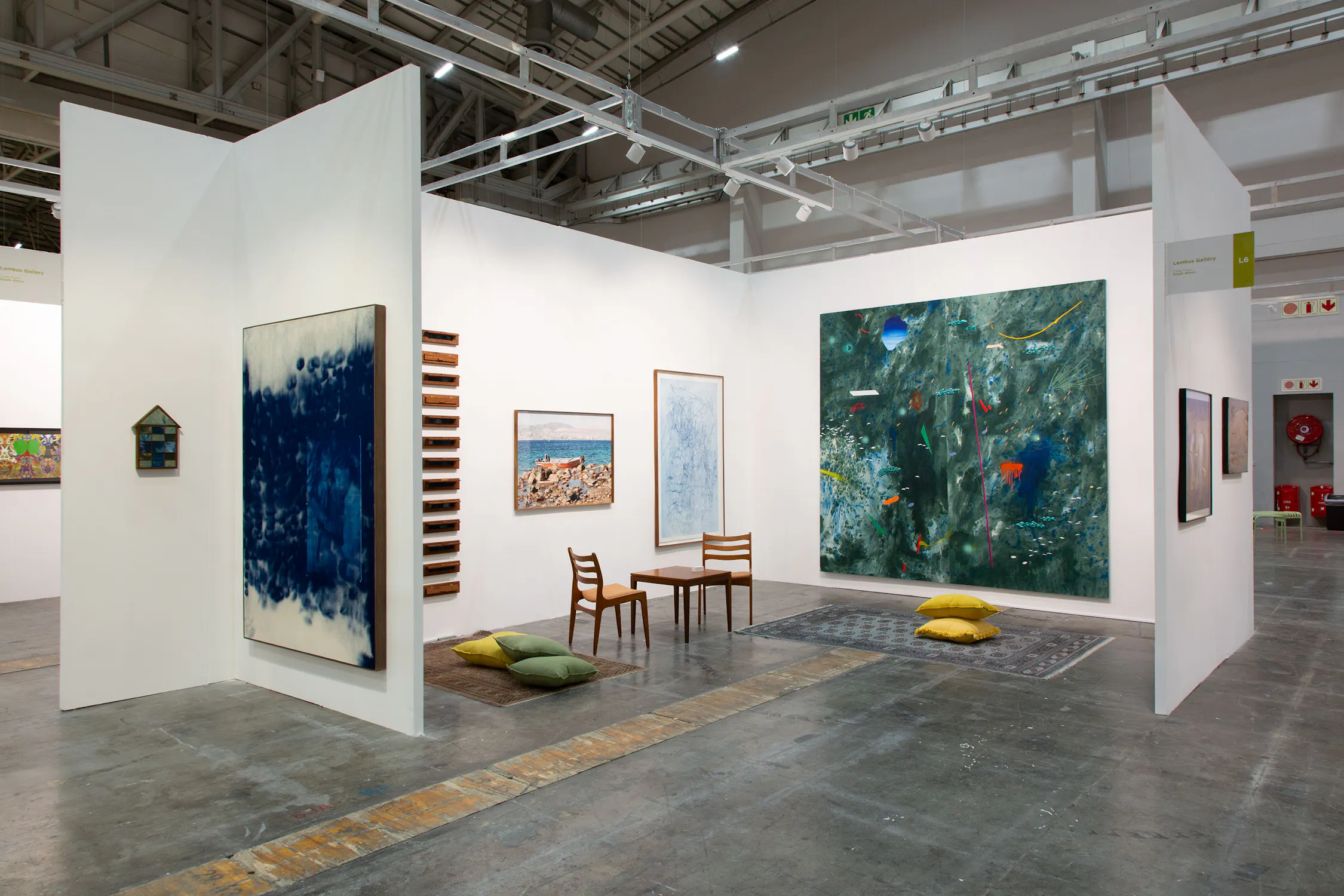 A contemporary art gallery with white walls displays abstract and landscape paintings. Two wooden chairs and several cushions sit on a small rug in the centre, inviting visitors to relax and view the artwork on concrete floors under industrial lighting.