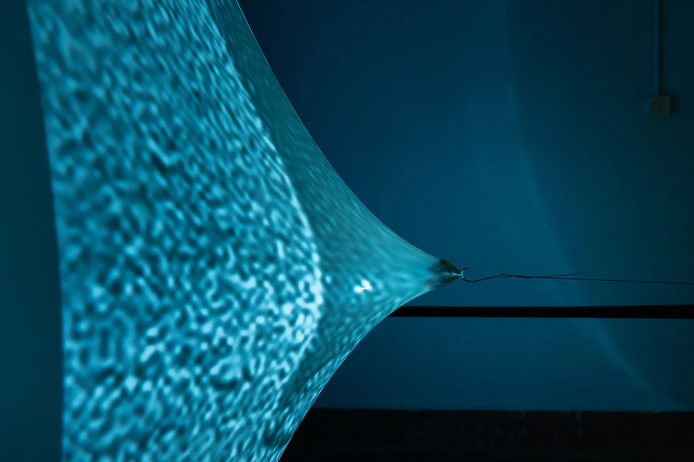 A taut, translucent blue sheet is stretched to a sharp point by a metal hook and thin wire against a dark background. The fabric displays a rippled, water-like light pattern.