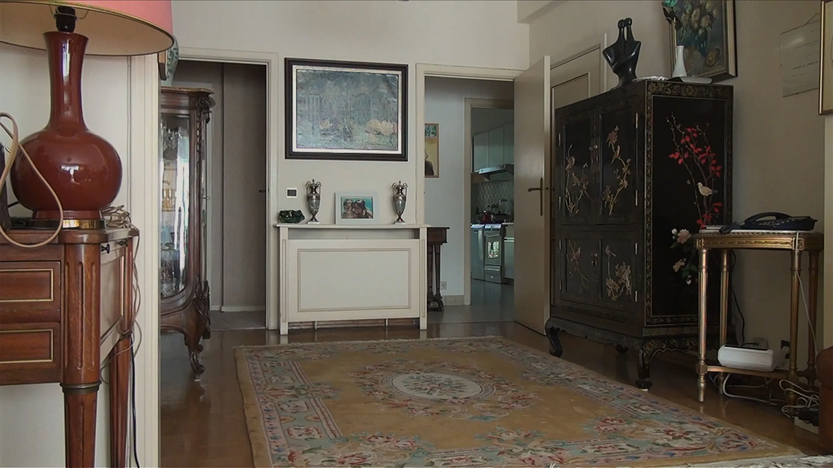 A furnished living room features a beige floral rug on wooden floors, leading towards doorways that reveal a hallway and kitchen. To the left is a wooden table with a large red lamp, and to the right stands a tall, dark, ornate cupboard decorated with painted floral motifs.