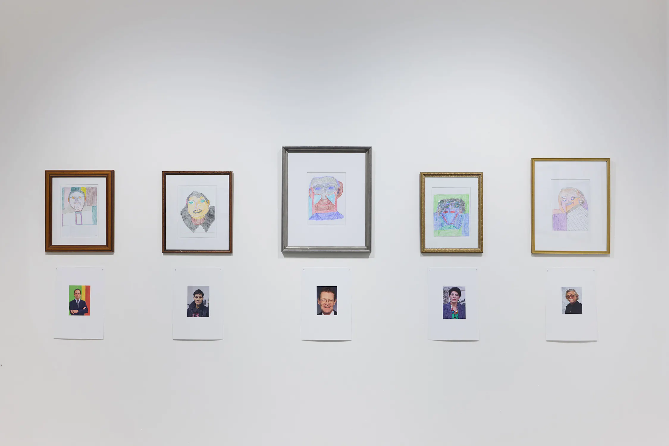 Five framed hand-drawn portraits hang on a white wall, each above a printed photo of a person, creating pairs. The portraits are colourful and abstract, while the photos appear to be more realistic, showing different individuals.