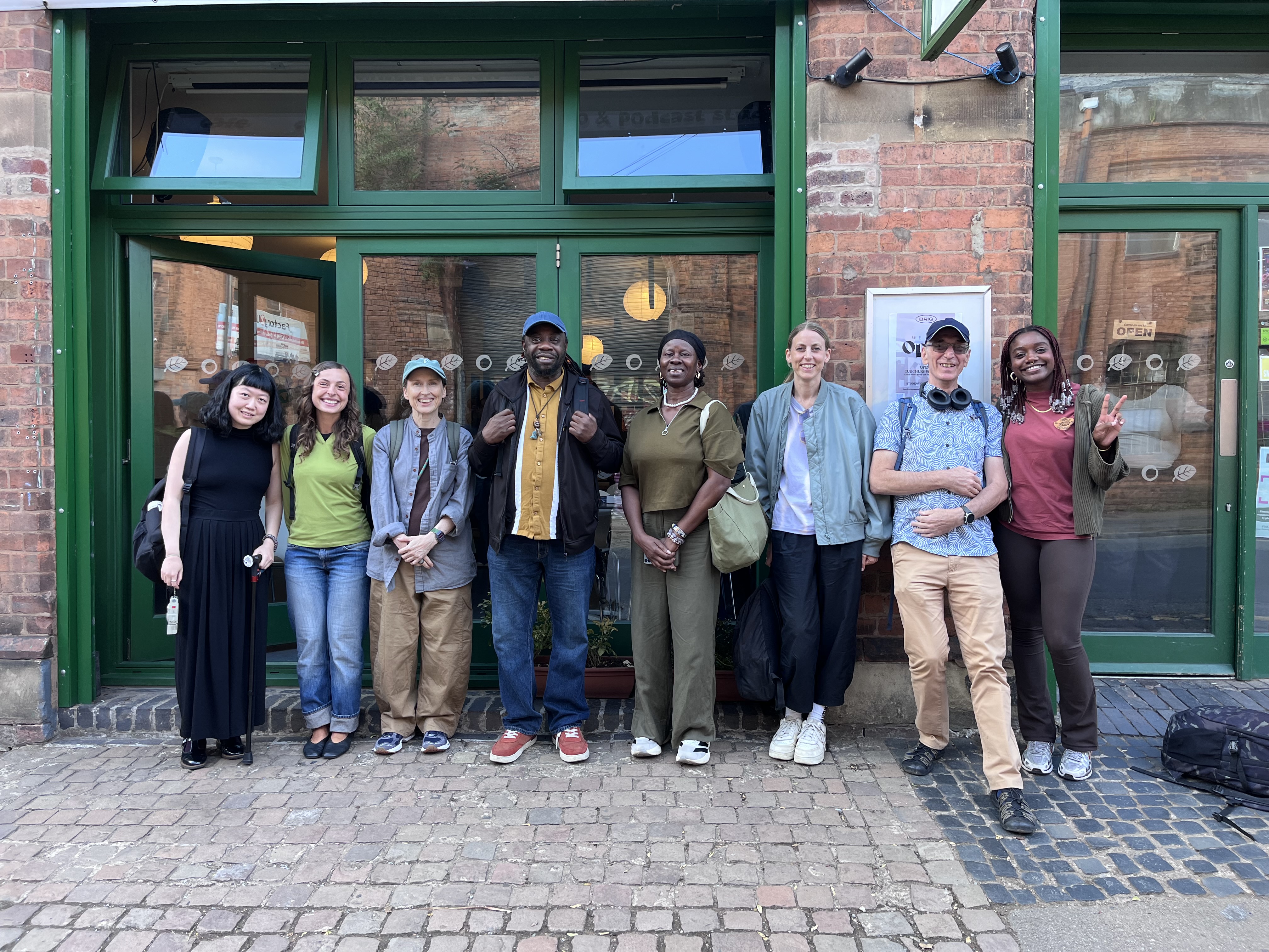 Eight people stand smiling in a row in front of a brick building with green trim and large windows. They wear casual clothes and have rucksacks or bags. Some have arms round each other; one makes a peace sign. The background shows pavement, windows, and a door.
