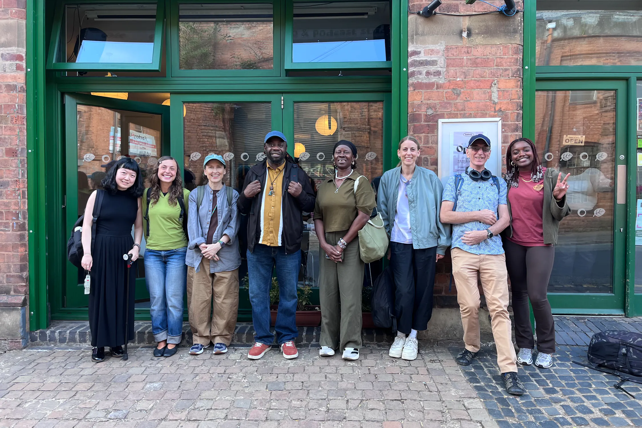 Eight people stand smiling in a row in front of a brick building with green trim and large windows. They wear casual clothes and have rucksacks or bags. Some have arms round each other; one makes a peace sign. The background shows pavement, windows, and a door.