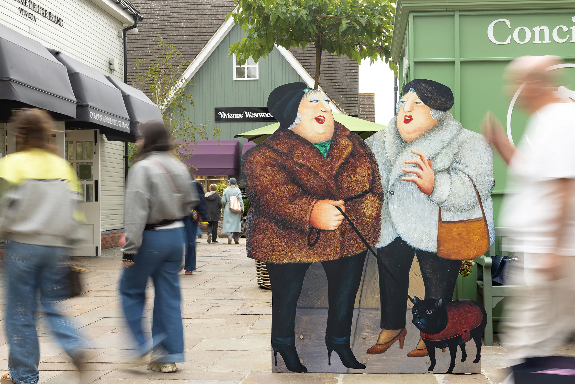A painted cut-out of two women in fur coats, one holding a lead with a small black dog, stands on a busy outdoor shopping street. Blurred people walk by, with shops and a green concierge kiosk in the background.