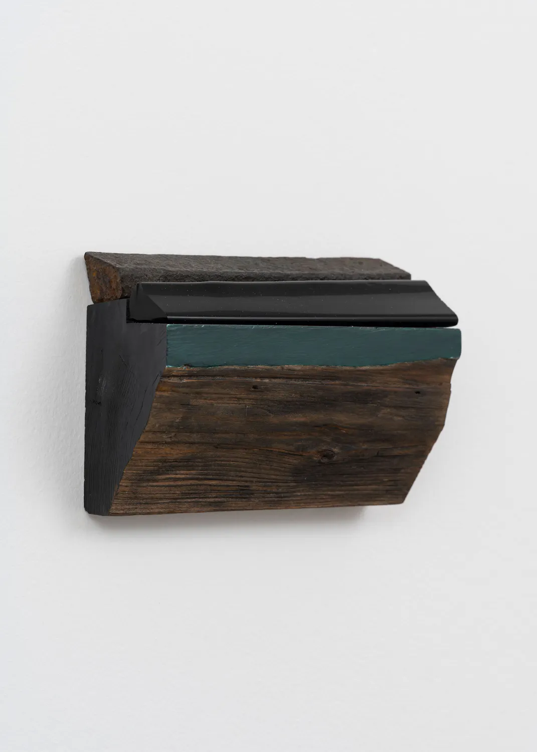 A small, multi-layered wooden sculpture mounted on a plain white wall. The piece features a rough, dark brown wooden base topped with a band of teal paint, a smooth black angled ledge, and a jagged, rusty metal top edge.
