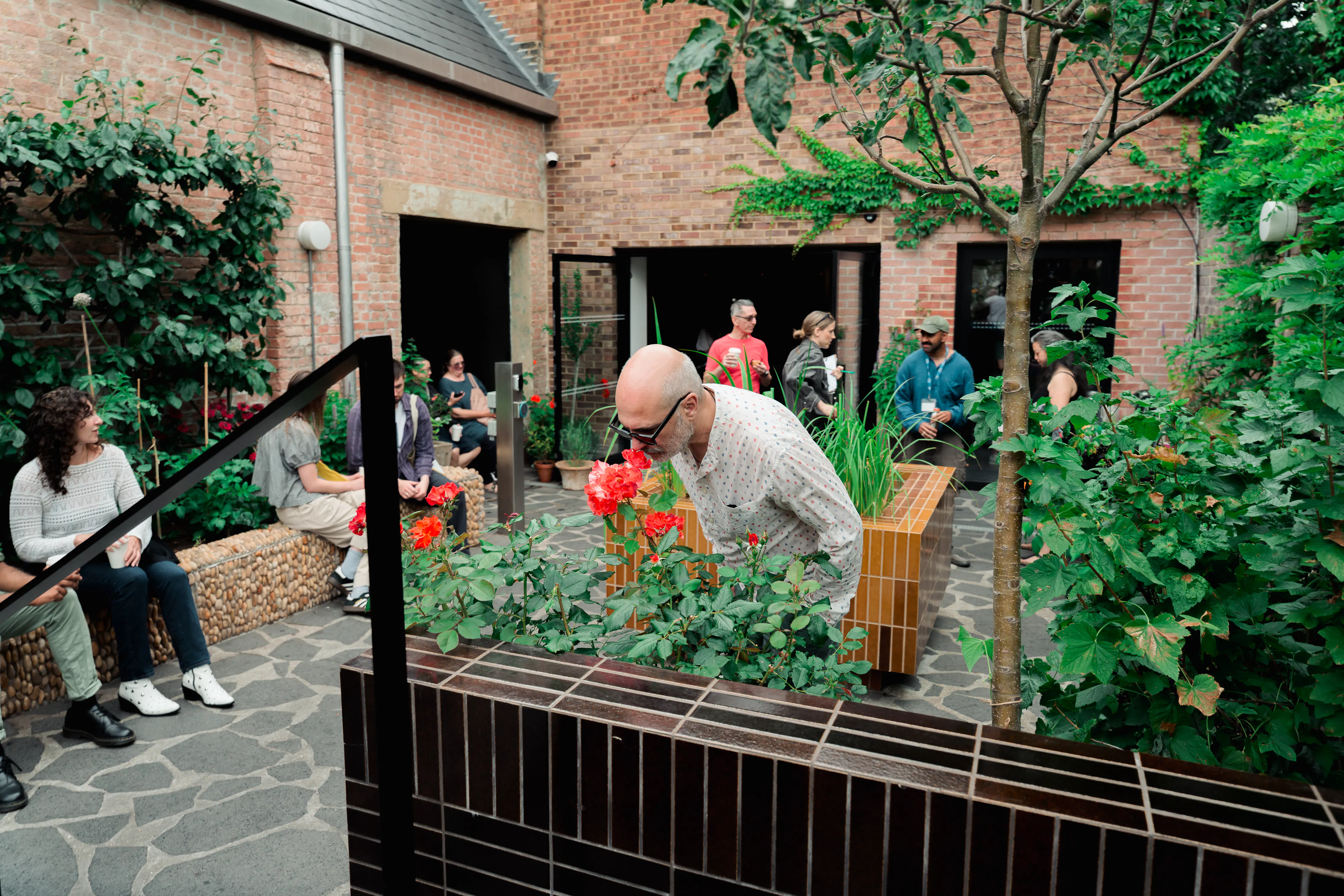 A bald man in a patterned shirt leans in to smell a red rose in a brick-walled courtyard. Nearby, other people sit on stone benches or stand and talk amongst various plants and trees.