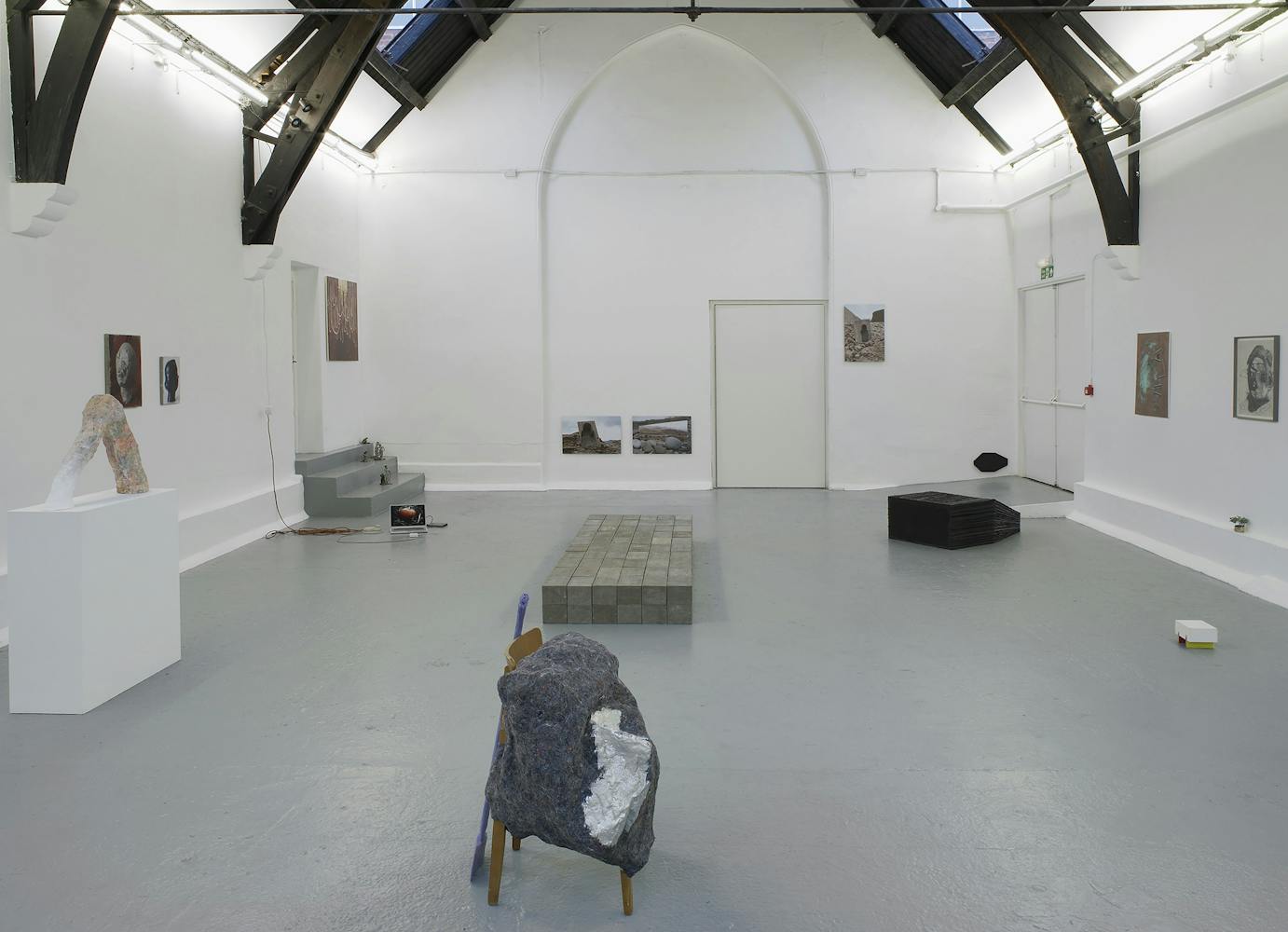 A contemporary art gallery with white walls and high, vaulted ceilings displays sculptures, paintings, and photos. Artworks are placed on the floor, plinths, and walls, in a minimalist, spacious arrangement.