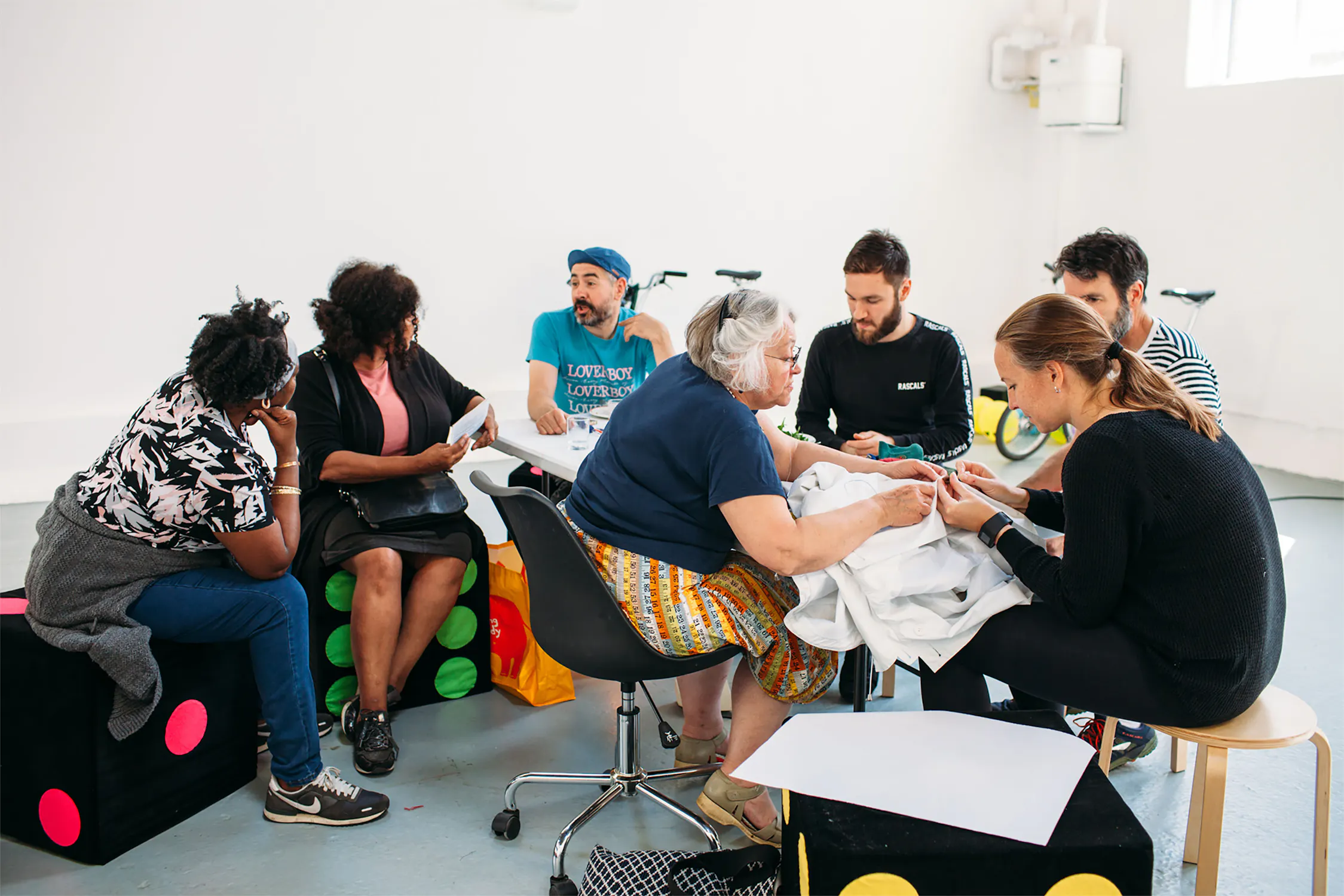 Seven adults sit in a circle on stools and cube seats in a bright room, engaged in a group activity involving fabric. They are talking, writing and interacting with materials, suggesting a collaborative or creative workshop.