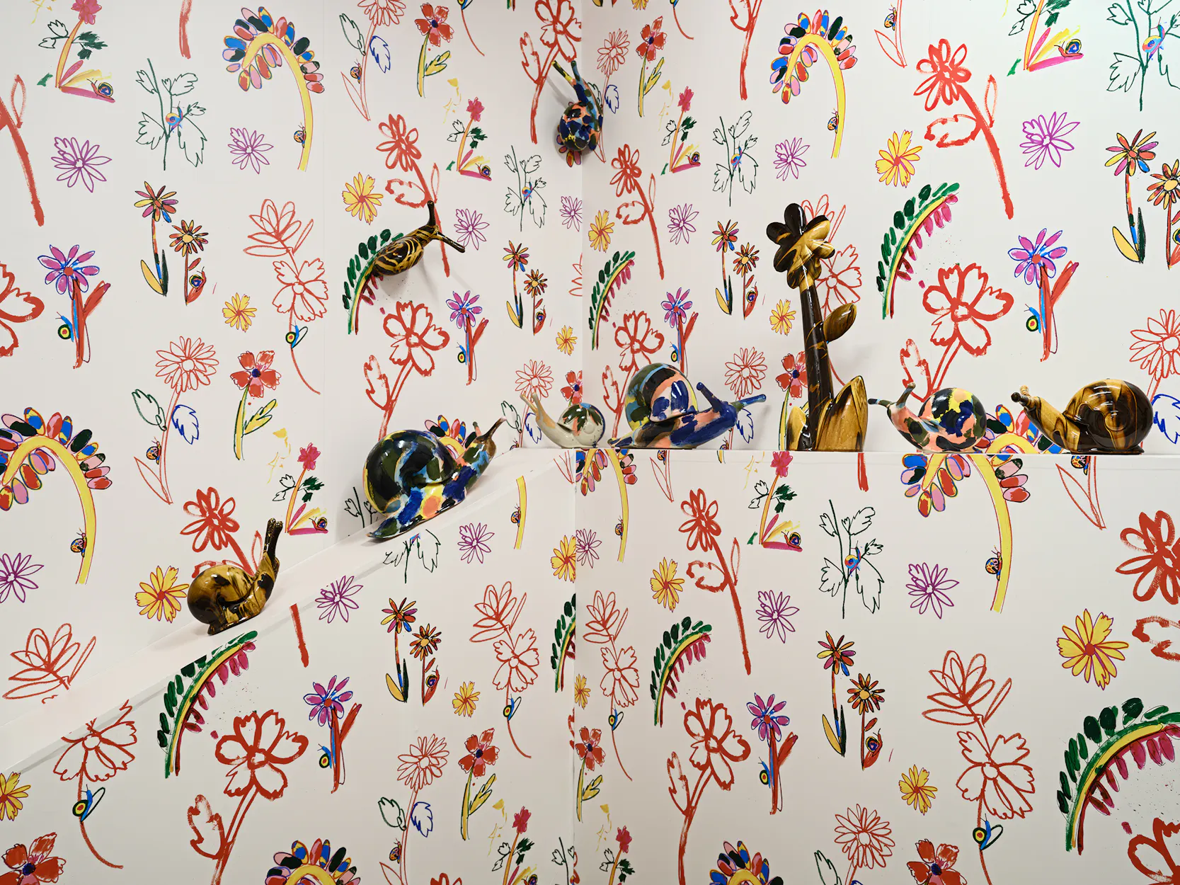 Colourful, whimsical floral and plant designs cover the walls and shelves of a room. Several glazed ceramic snail sculptures are displayed on the angled white shelves, blending with the lively botanical wallpaper.