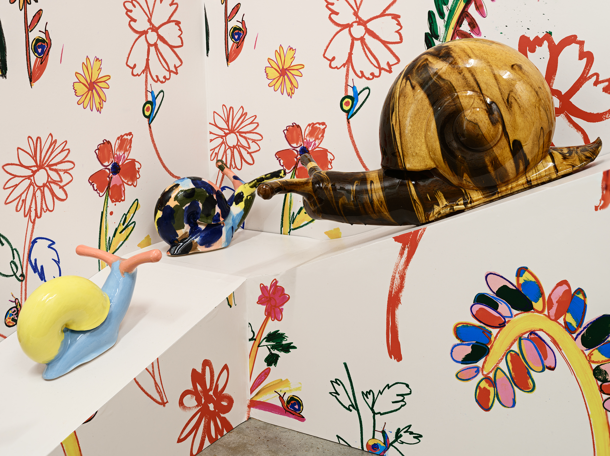 Three colourful ceramic snail sculptures sit on a white ramp against a wall covered in playful, hand-drawn red and yellow flowers and leaves.