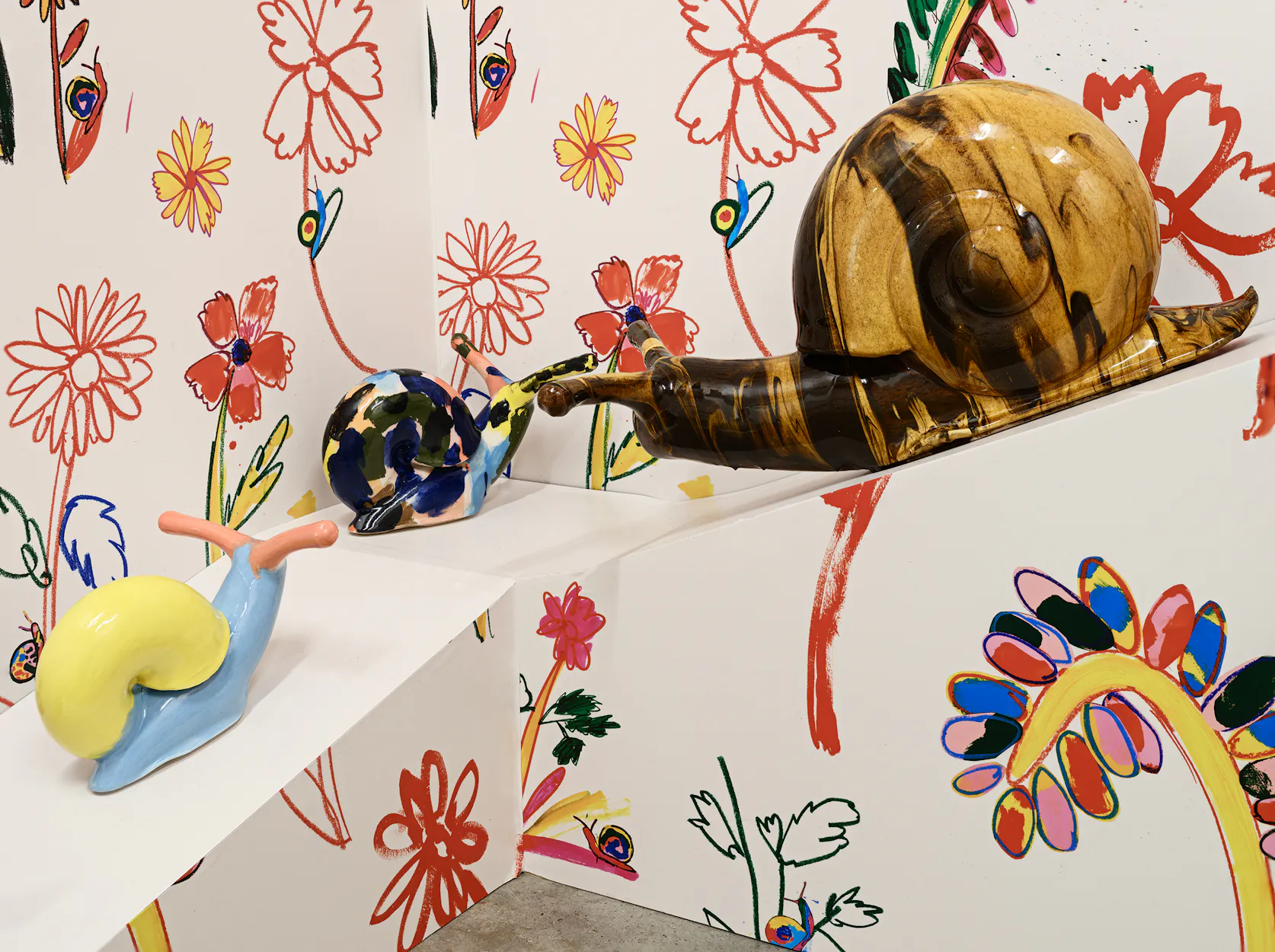 Three colourful ceramic snail sculptures sit on a white ramp against a wall covered in playful, hand-drawn red and yellow flowers and leaves.