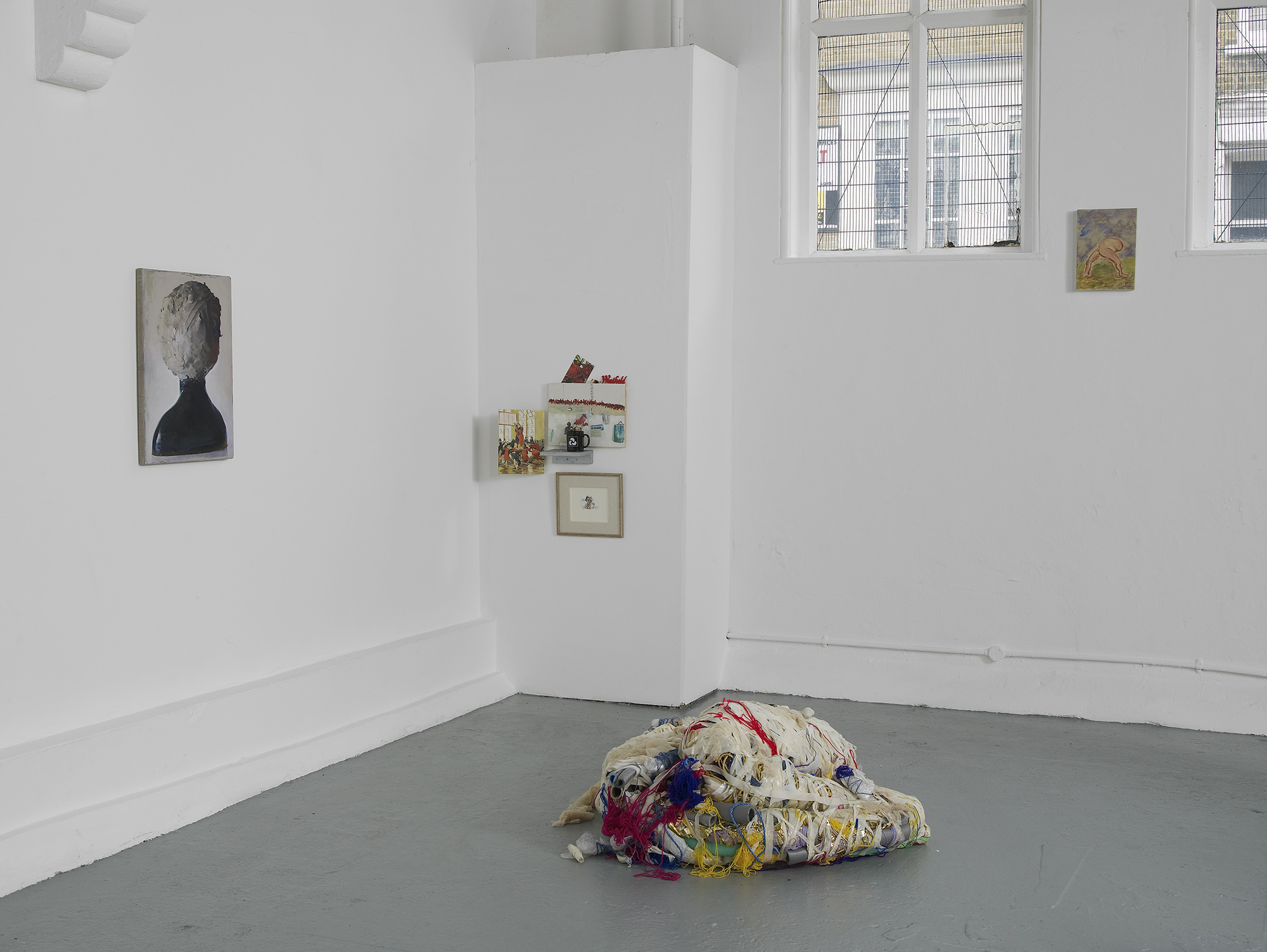 A contemporary art gallery with white walls displays small artworks and photographs, while a colorful, textured fabric sculpture sits on the gray floor in the center of the room. Sunlight enters through a window.