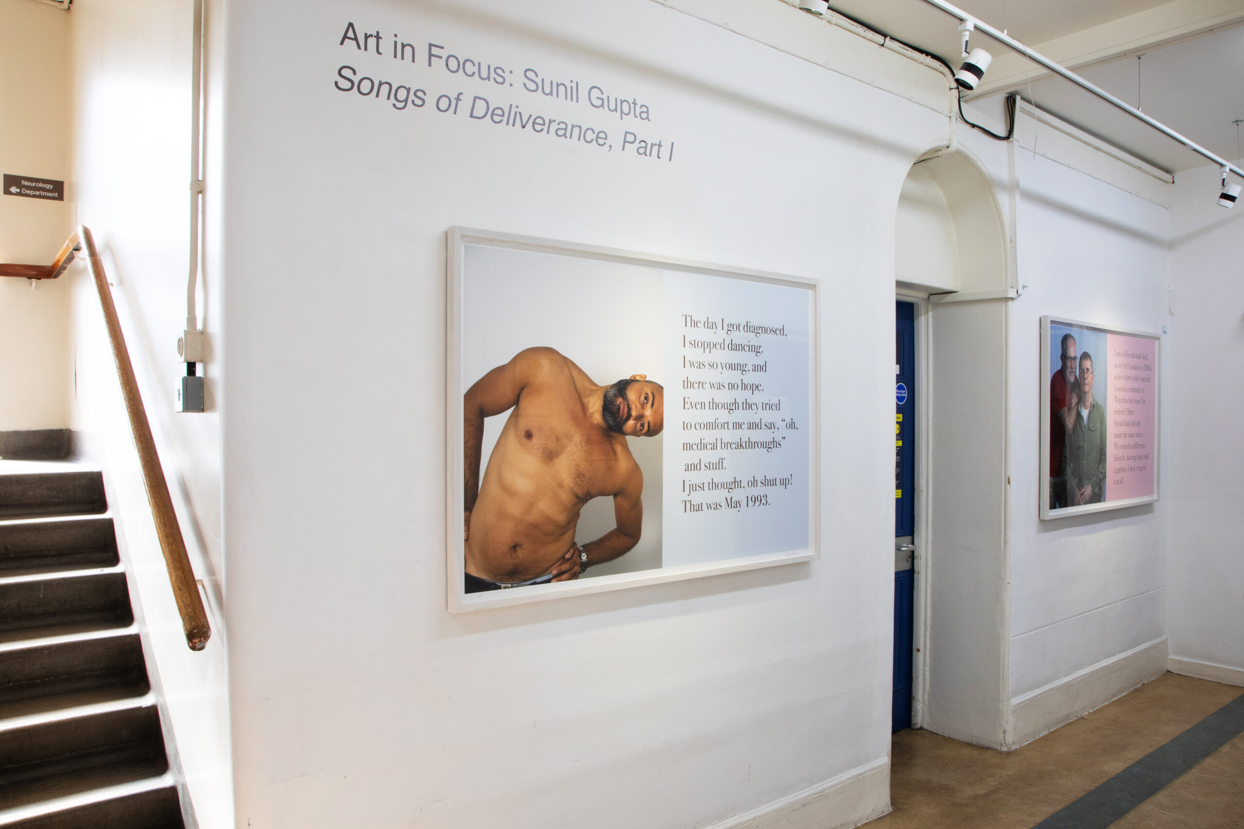 An artwork featuring a photograph of a shirtless man with a hand on his hip and accompanying text is displayed in a hospital corridor. Stairs are visible on the left.