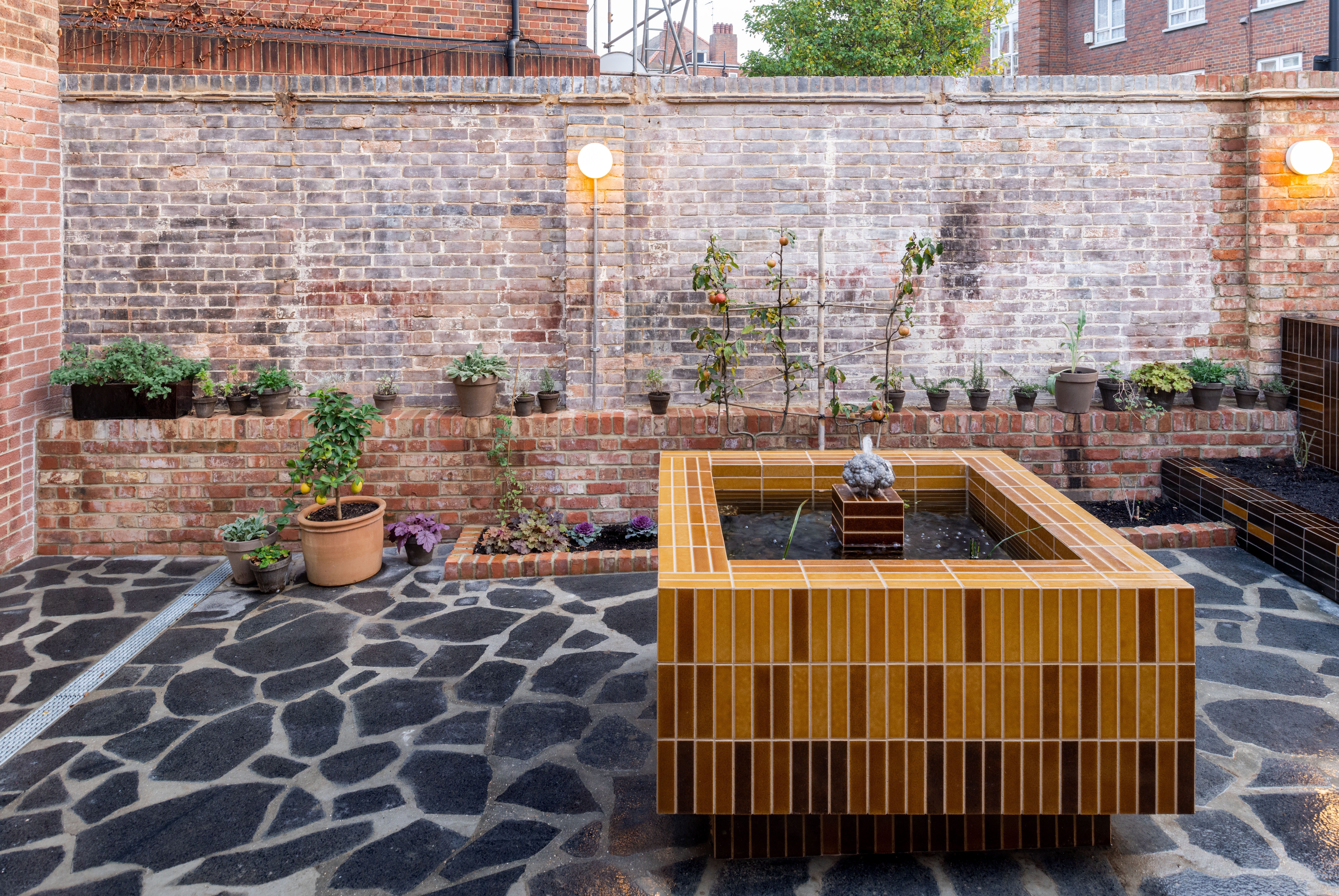 A small urban garden with a tiled water feature in the centre, surrounded by various potted plants on a brick ledge against a brick wall. The area has a stone-paved floor and is illuminated by two wall-mounted lights.