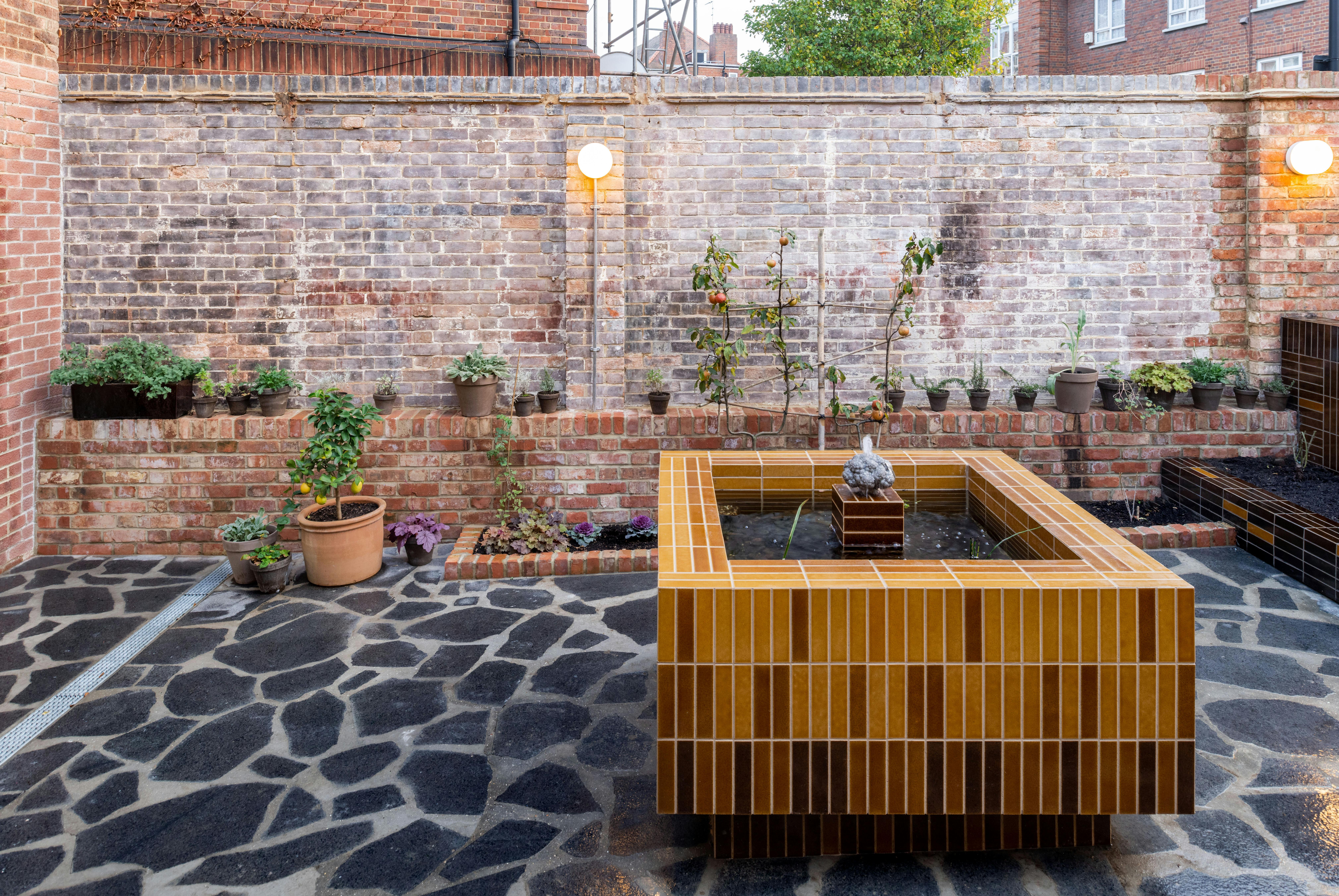 A small urban garden with a tiled water feature in the centre, surrounded by various potted plants on a brick ledge against a brick wall. The area has a stone-paved floor and is illuminated by two wall-mounted lights.