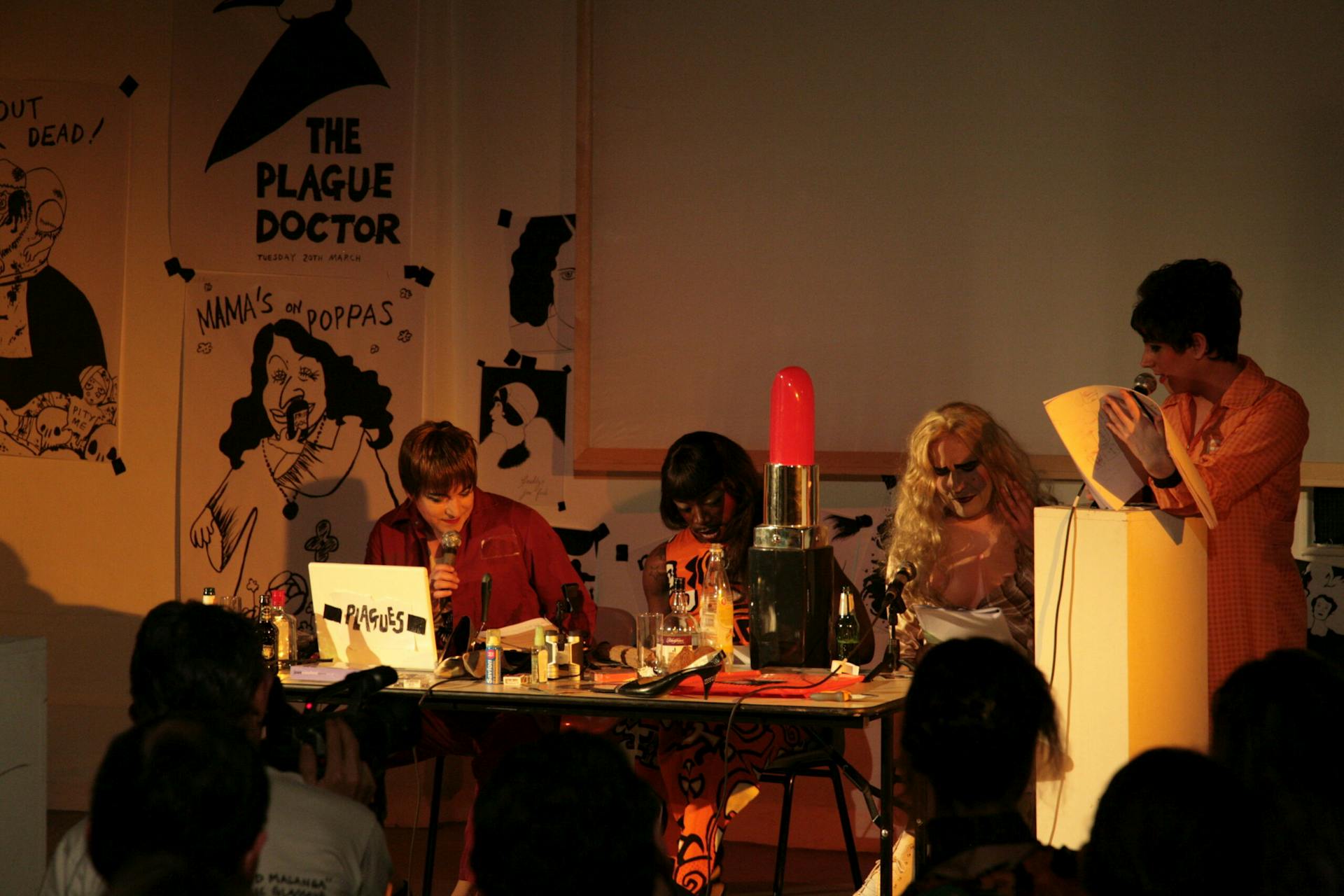 Four costumed performers sit at a table in front of an audience. The table has a giant novelty red lipstick, a high heel and a laptop. The wall in the background is plastered with black and white illustrated posters