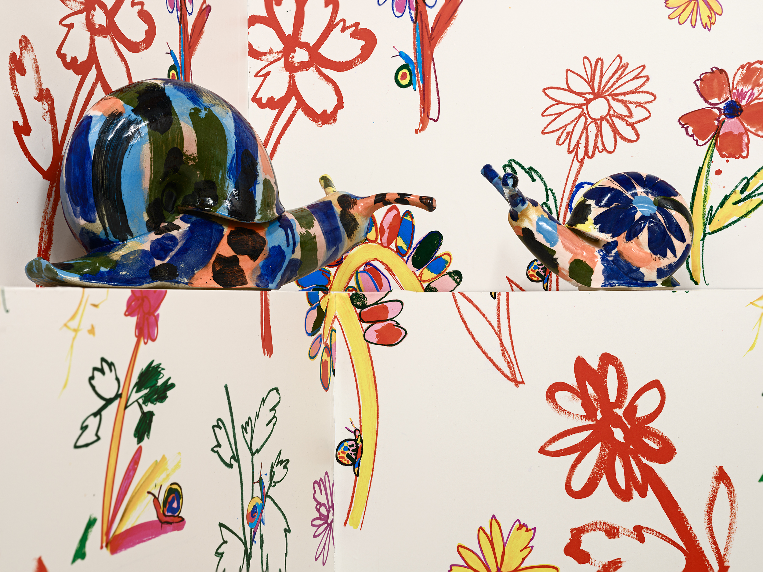 Two colourful ceramic snail sculptures by Supergroup are placed against a white background decorated with vibrant, hand-drawn floral and leaf patterns in red, orange, green, and blue. The snails face each other amid the lively designs.