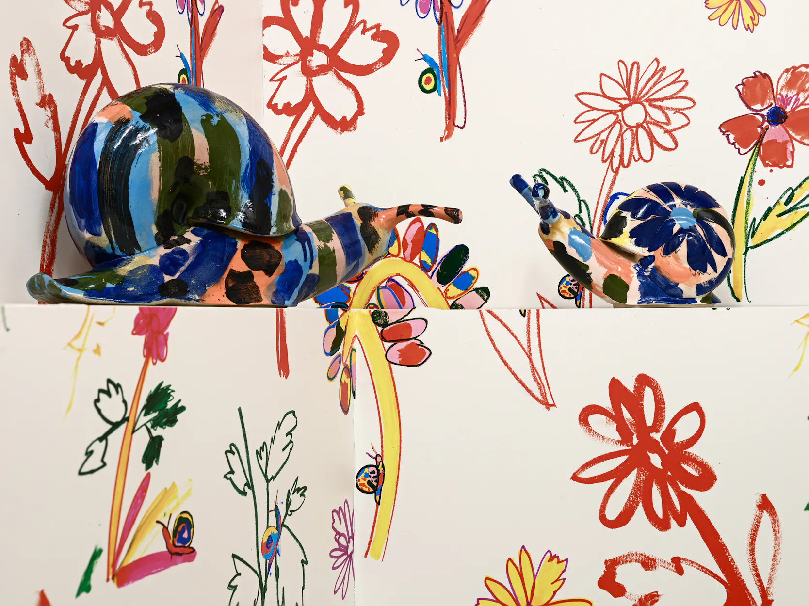 Two colourful ceramic snail sculptures by Supergroup are placed against a white background decorated with vibrant, hand-drawn floral and leaf patterns in red, orange, green, and blue. The snails face each other amid the lively designs.