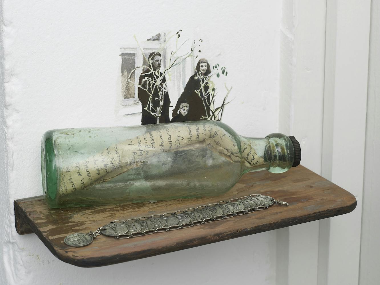 A glass bottle with a handwritten letter inside rests on a wooden shelf, next to a coin bracelet. Behind them is a wall painting of three people, including a child, holding thin branches.