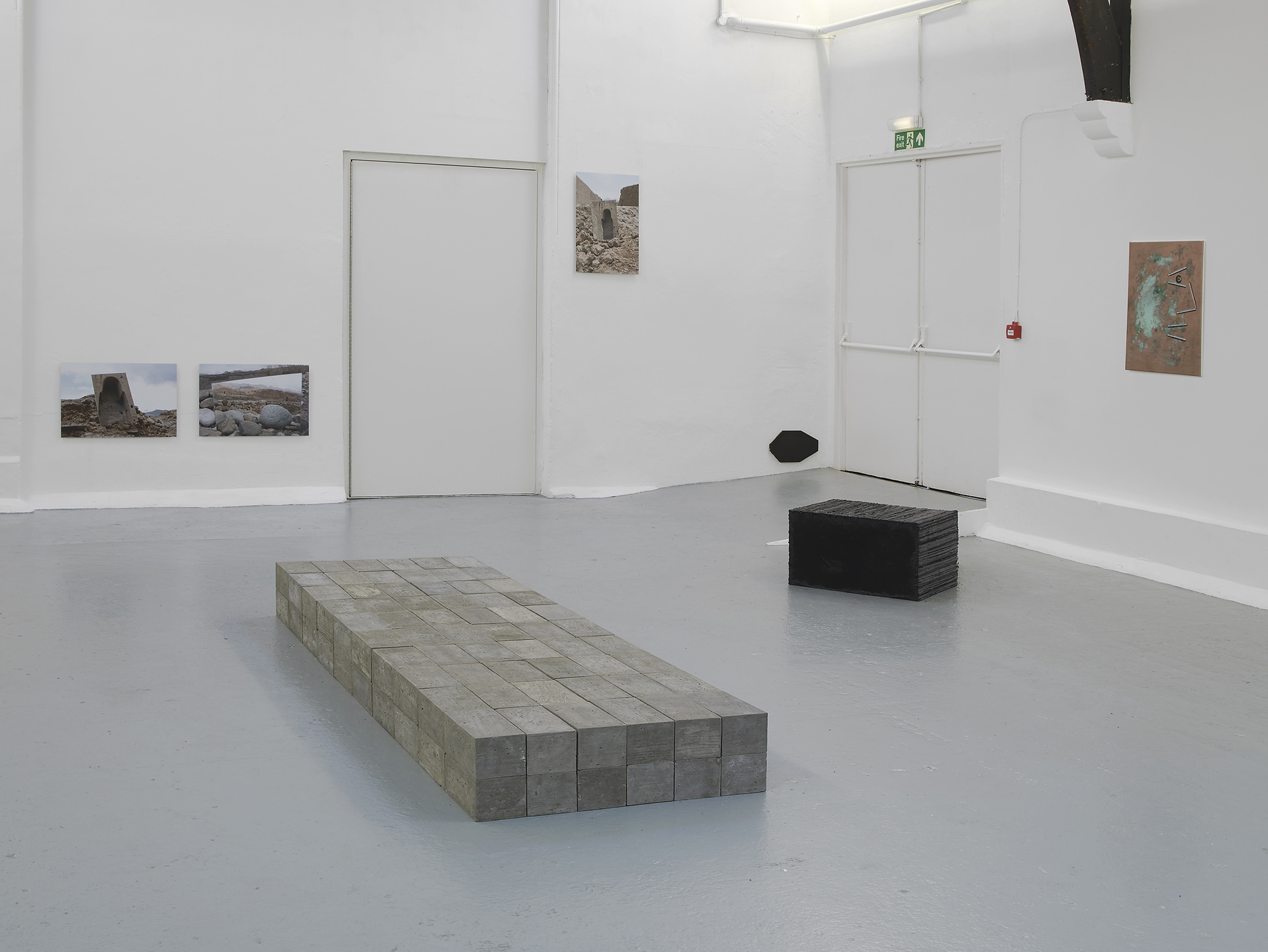 A contemporary art gallery with white walls and high, vaulted ceilings displays sculptures, paintings, and photos. Artworks are placed on the floor, plinths, and walls, in a minimalist, spacious arrangement.