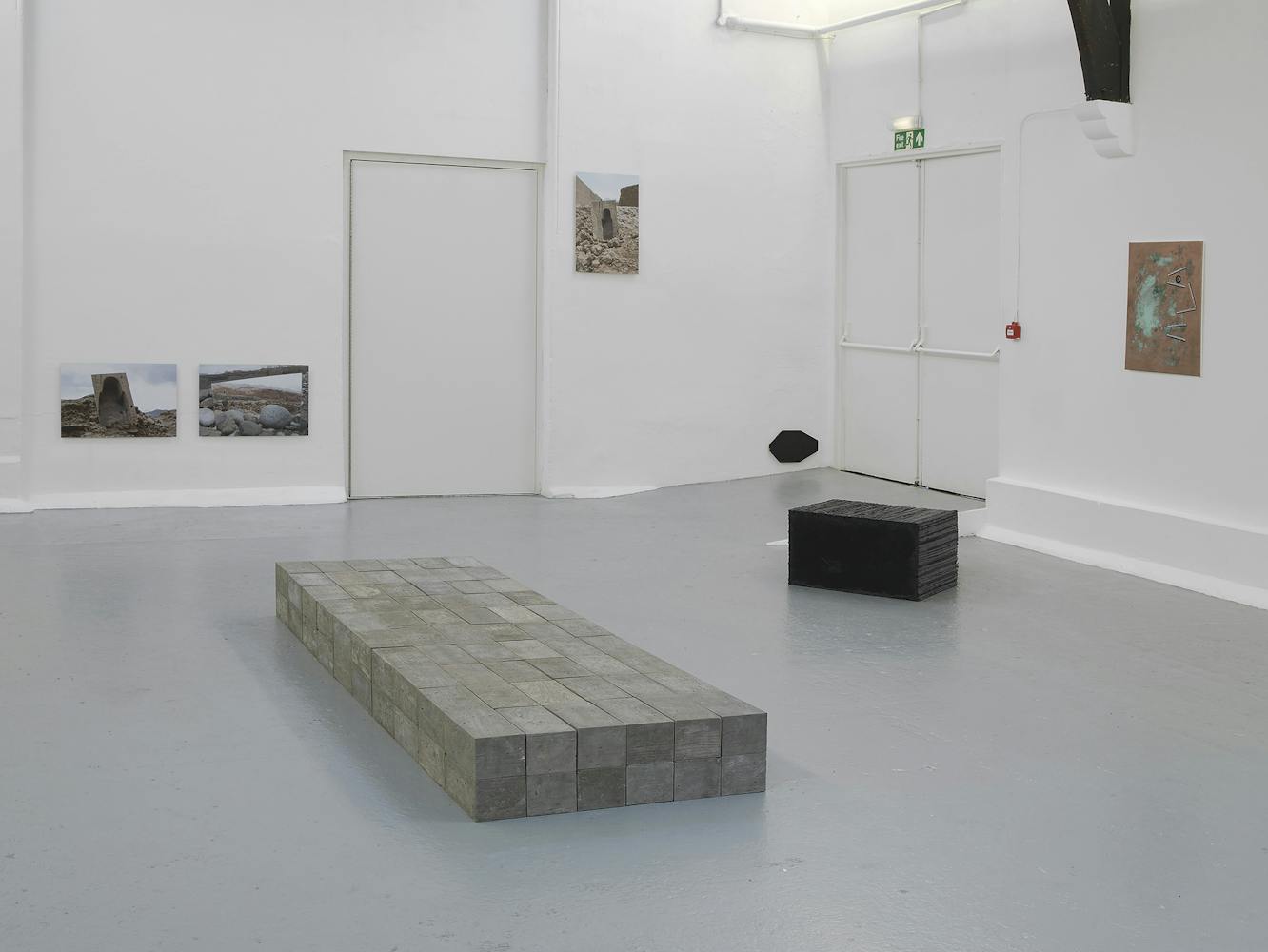 A contemporary art gallery with white walls and high, vaulted ceilings displays sculptures, paintings, and photos. Artworks are placed on the floor, plinths, and walls, in a minimalist, spacious arrangement.