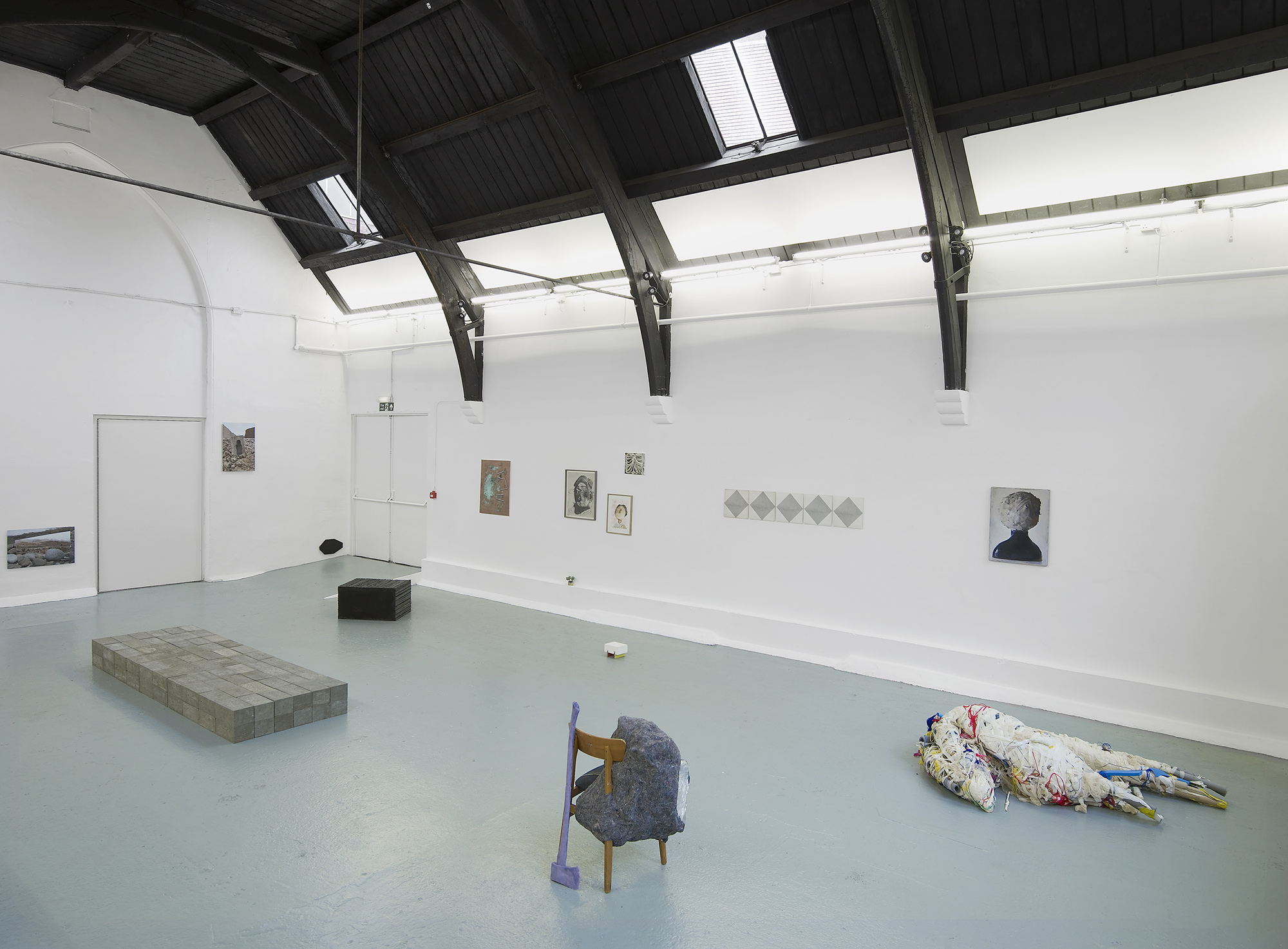 A contemporary art gallery with white walls and black ceiling beams displays various artworks, including paintings on the walls, a brick platform, a small black block, a chair with fabric, and a fabric-wrapped figure on the floor.