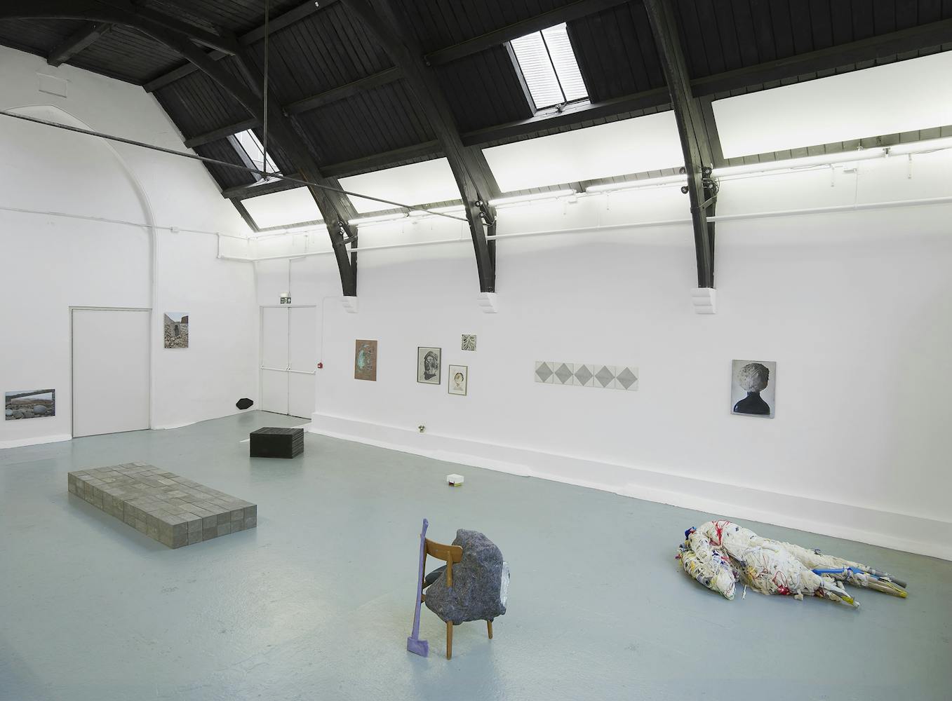 A contemporary art gallery with white walls and black ceiling beams displays various artworks, including paintings on the walls, a brick platform, a small black block, a chair with fabric, and a fabric-wrapped figure on the floor.