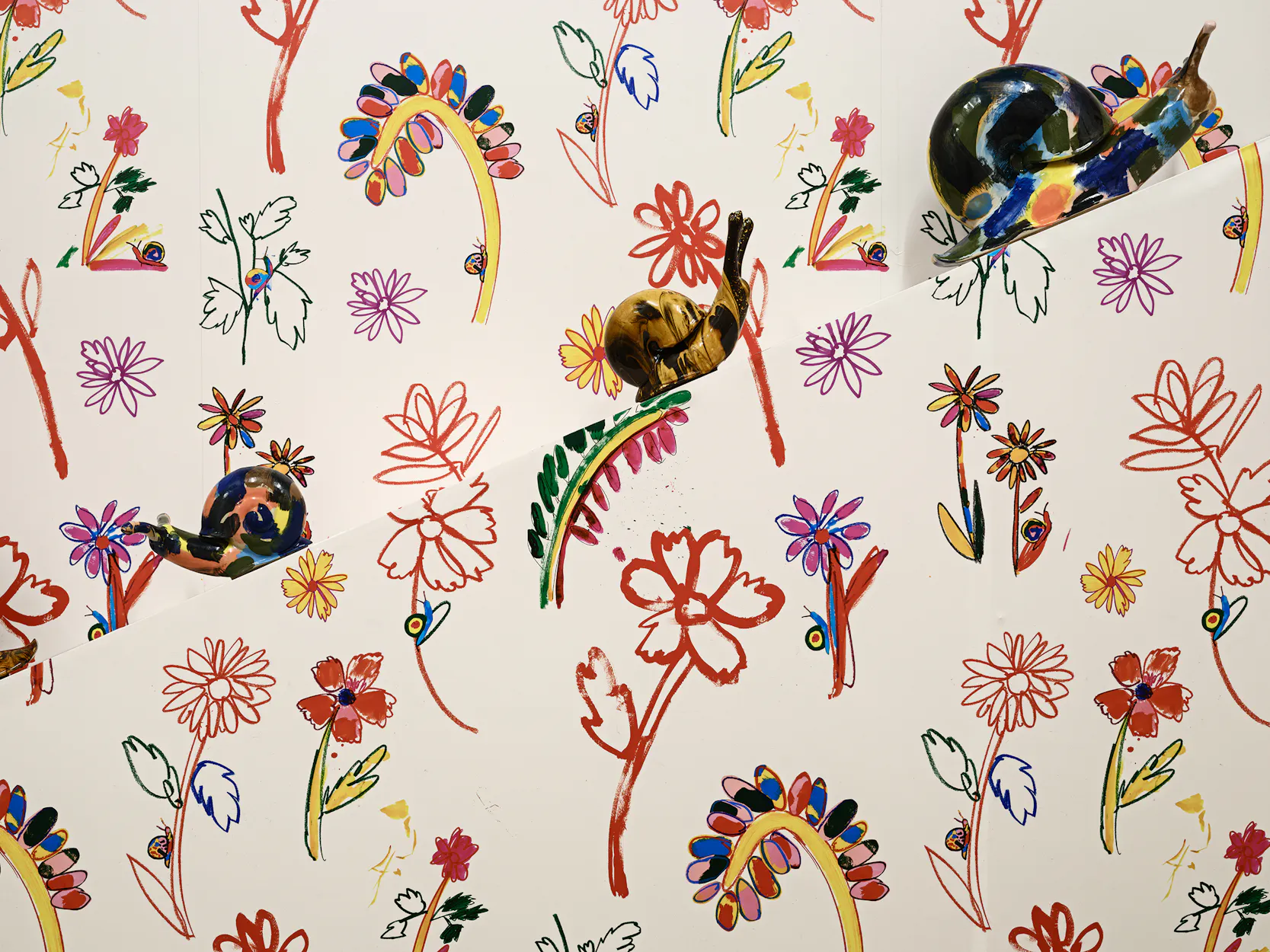 Colourful ceramic snails by Supergroup are placed on a wall covered with a playful, hand-drawn floral pattern featuring various red, yellow, green, and purple flowers and leaves on a white background.