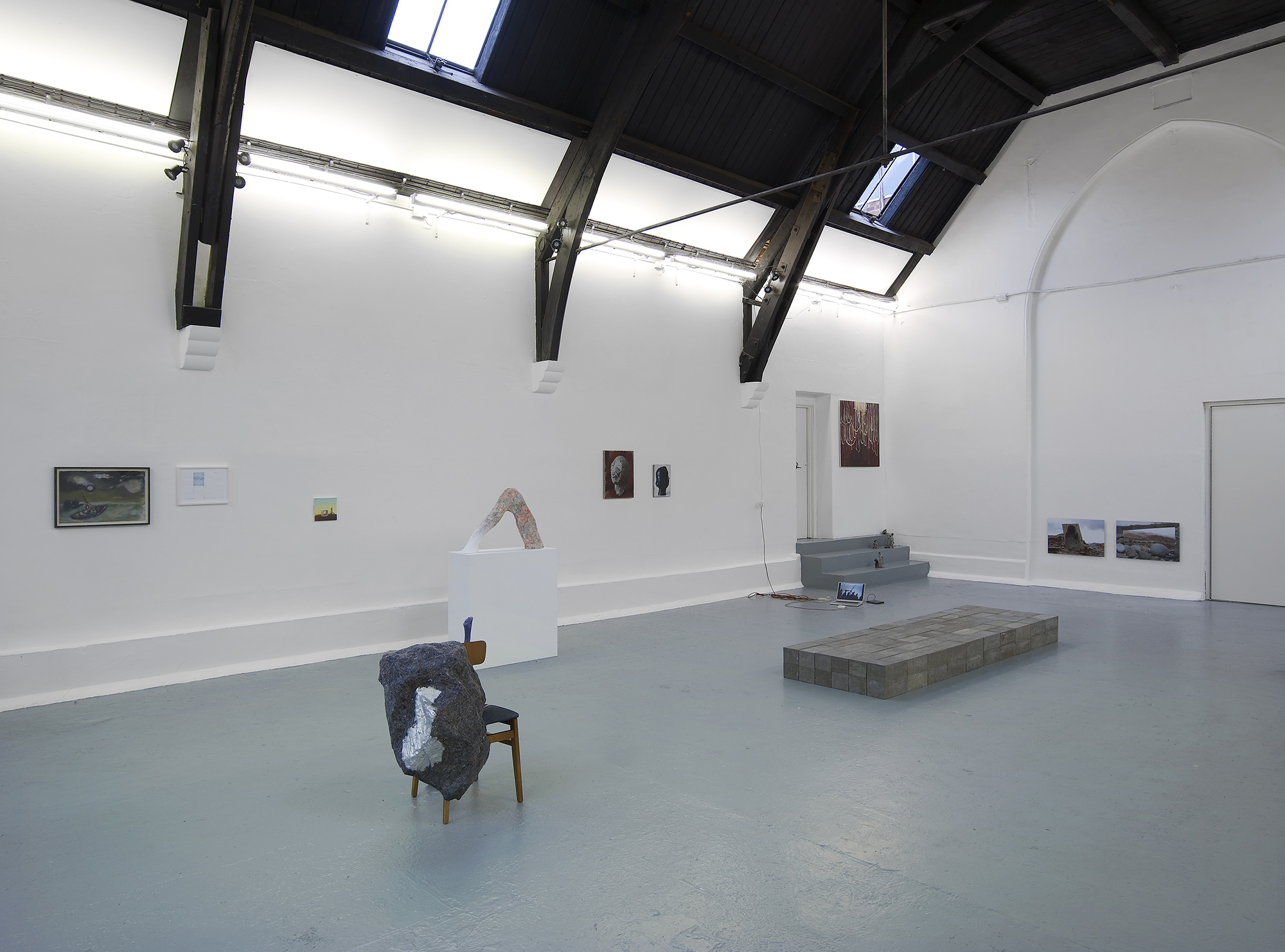 A contemporary art gallery with high, arched ceilings, white walls, and grey floors, featuring various artworks including paintings, a sculpture on a pedestal, a chair with fabric, and low platforms.