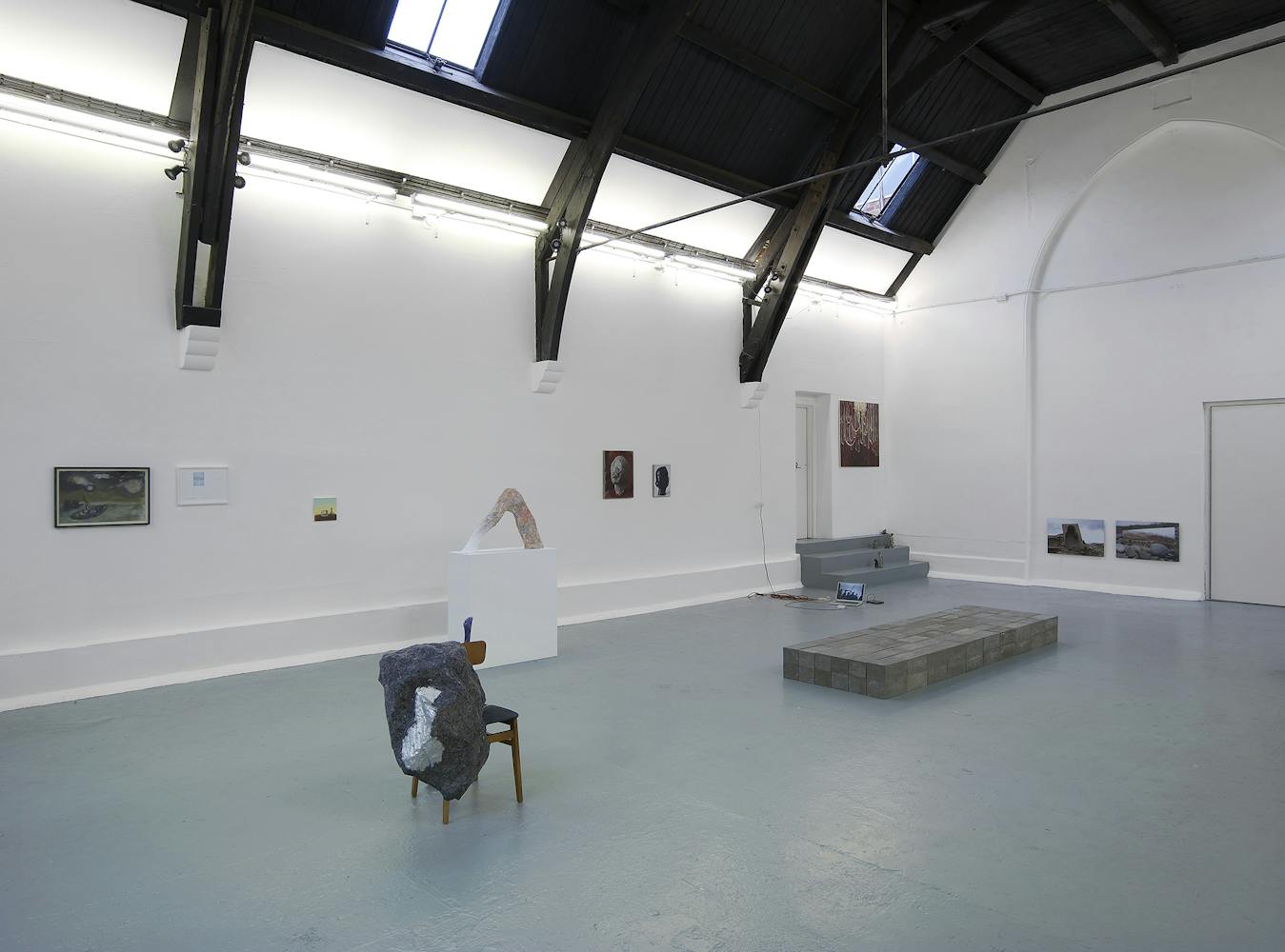 A contemporary art gallery with high, arched ceilings, white walls, and grey floors, featuring various artworks including paintings, a sculpture on a pedestal, a chair with fabric, and low platforms.