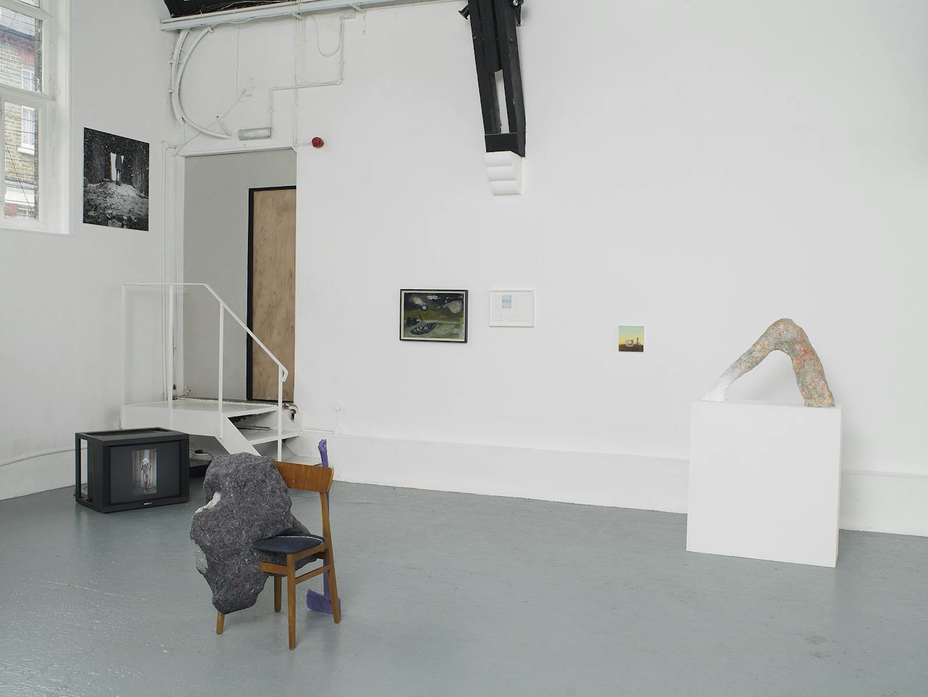 A contemporary art gallery with white walls and high, vaulted ceilings displays sculptures, paintings, and photos. Artworks are placed on the floor, plinths, and walls, in a minimalist, spacious arrangement.