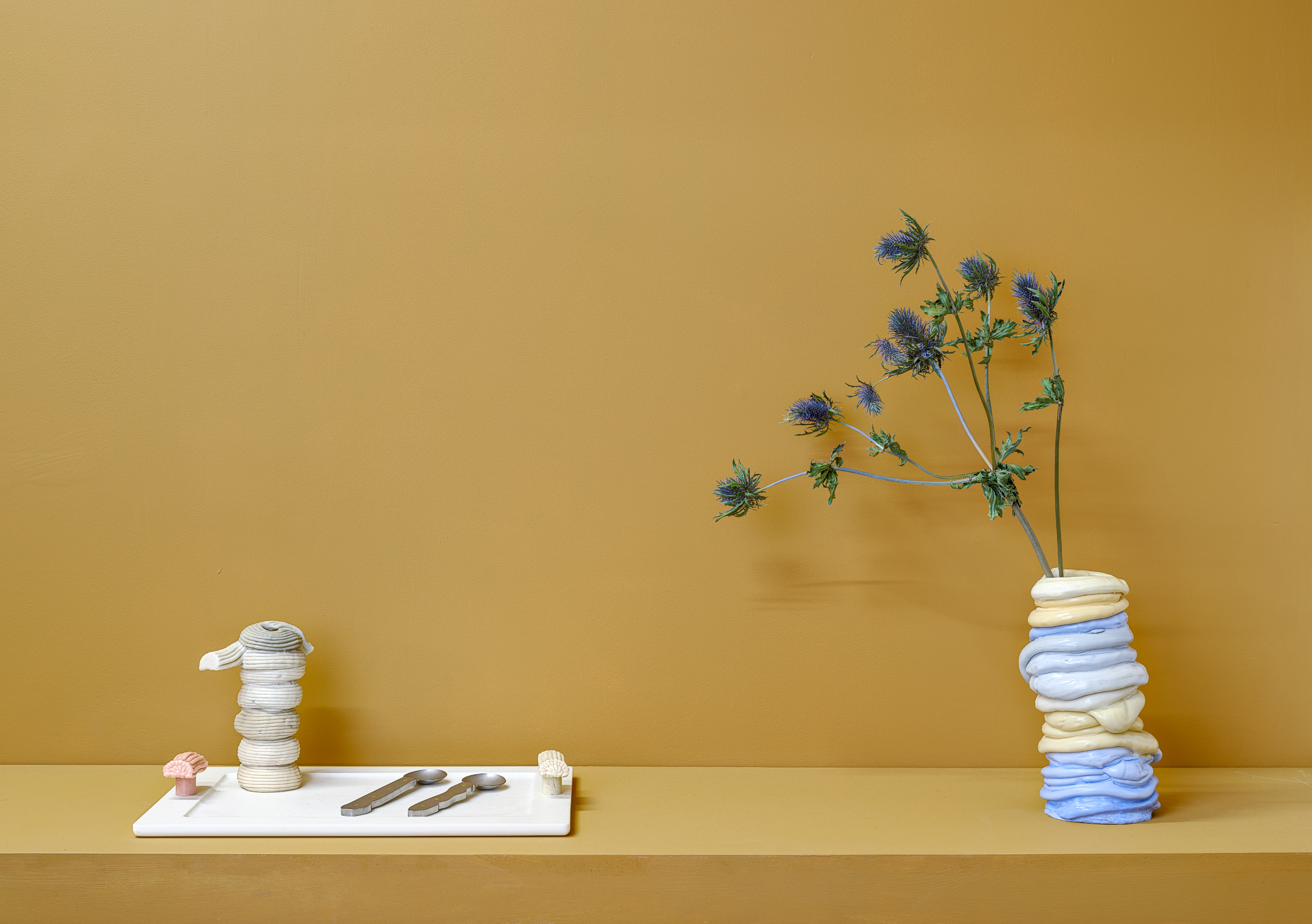 A minimalist display with a mustard yellow background featuring a pastel blue and yellow vase holding spiky purple flowers and a tray with stacked sculptural plastic vases and three metal spoons, all arranged on a light surface.