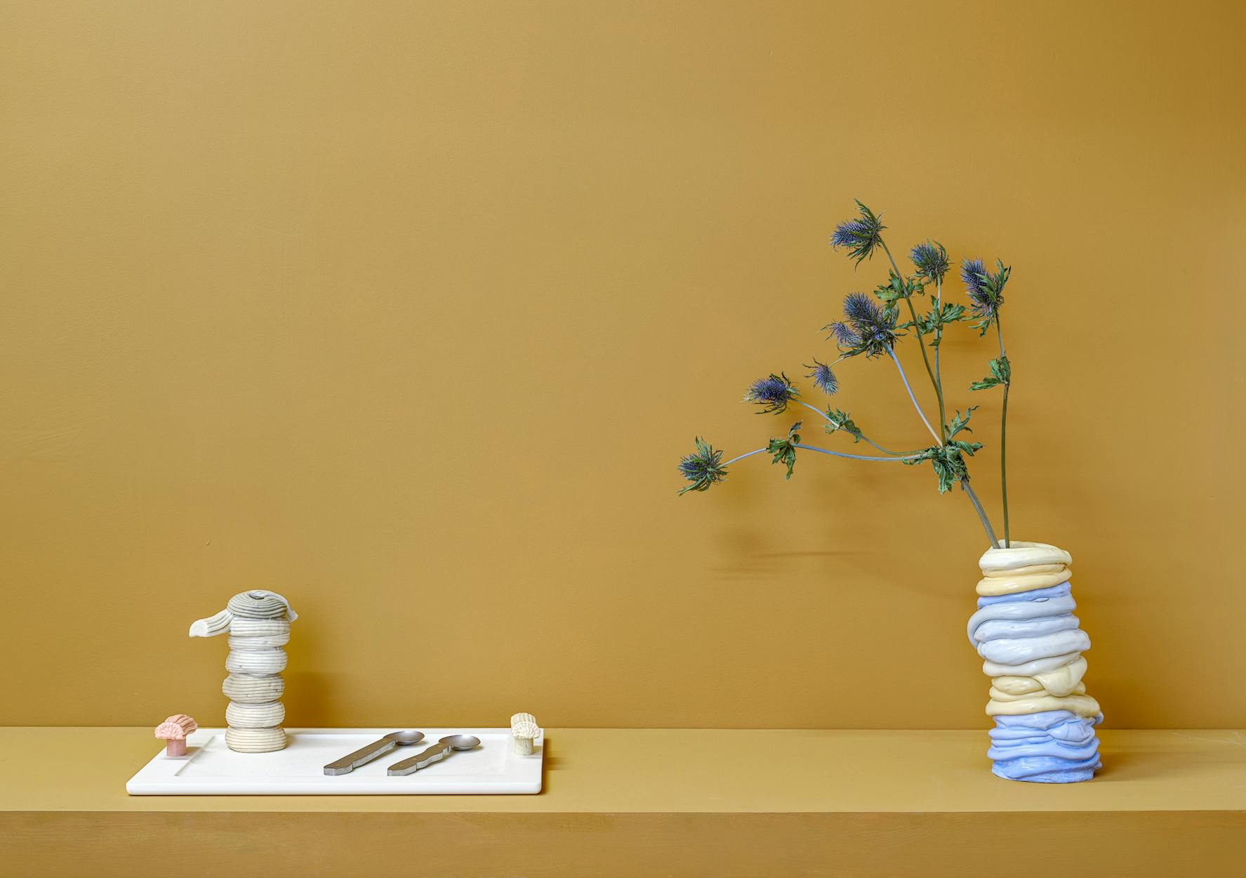 A minimalist display with a mustard yellow background featuring a pastel blue and yellow vase holding spiky purple flowers and a tray with stacked sculptural plastic vases and three metal spoons, all arranged on a light surface.