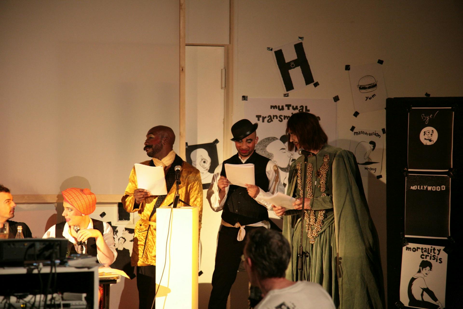 Three costumed performers stand in front of an audience. the performers are holding scripts.