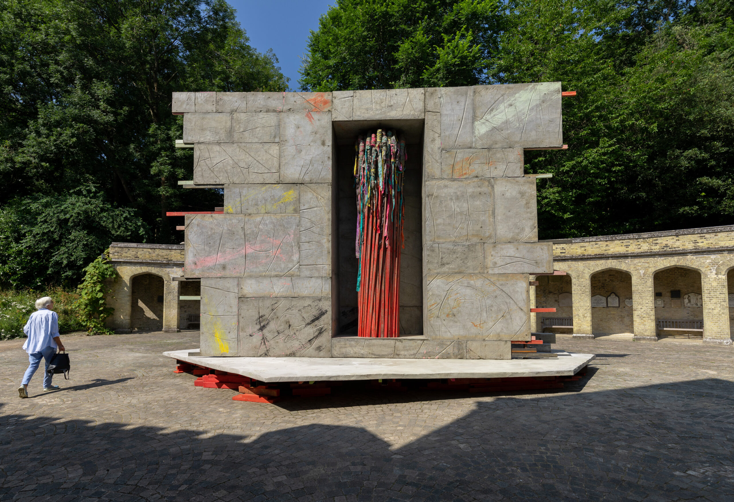 A person walks near a large outdoor sculpture made of rectangular stone slabs with a central vertical opening. Inside the opening, colourful poles stand upright. The installation is set in a paved courtyard surrounded by old brick walls and greenery.