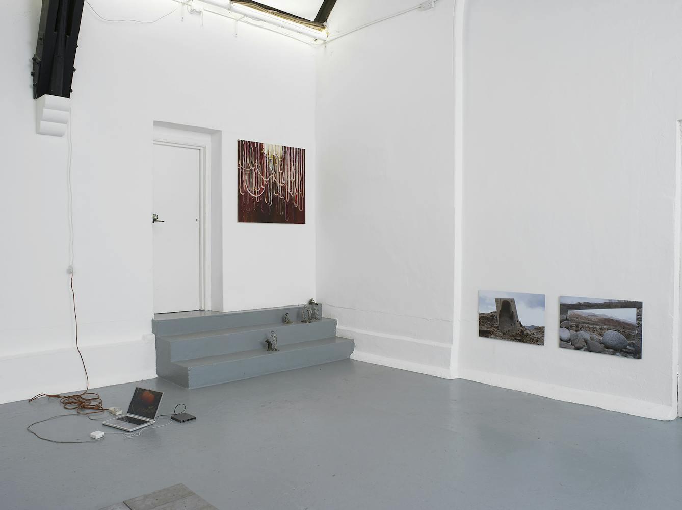 A contemporary art gallery with white walls and high, vaulted ceilings displays sculptures, paintings, and photos. Artworks are placed on the floor, plinths, and walls, in a minimalist, spacious arrangement.