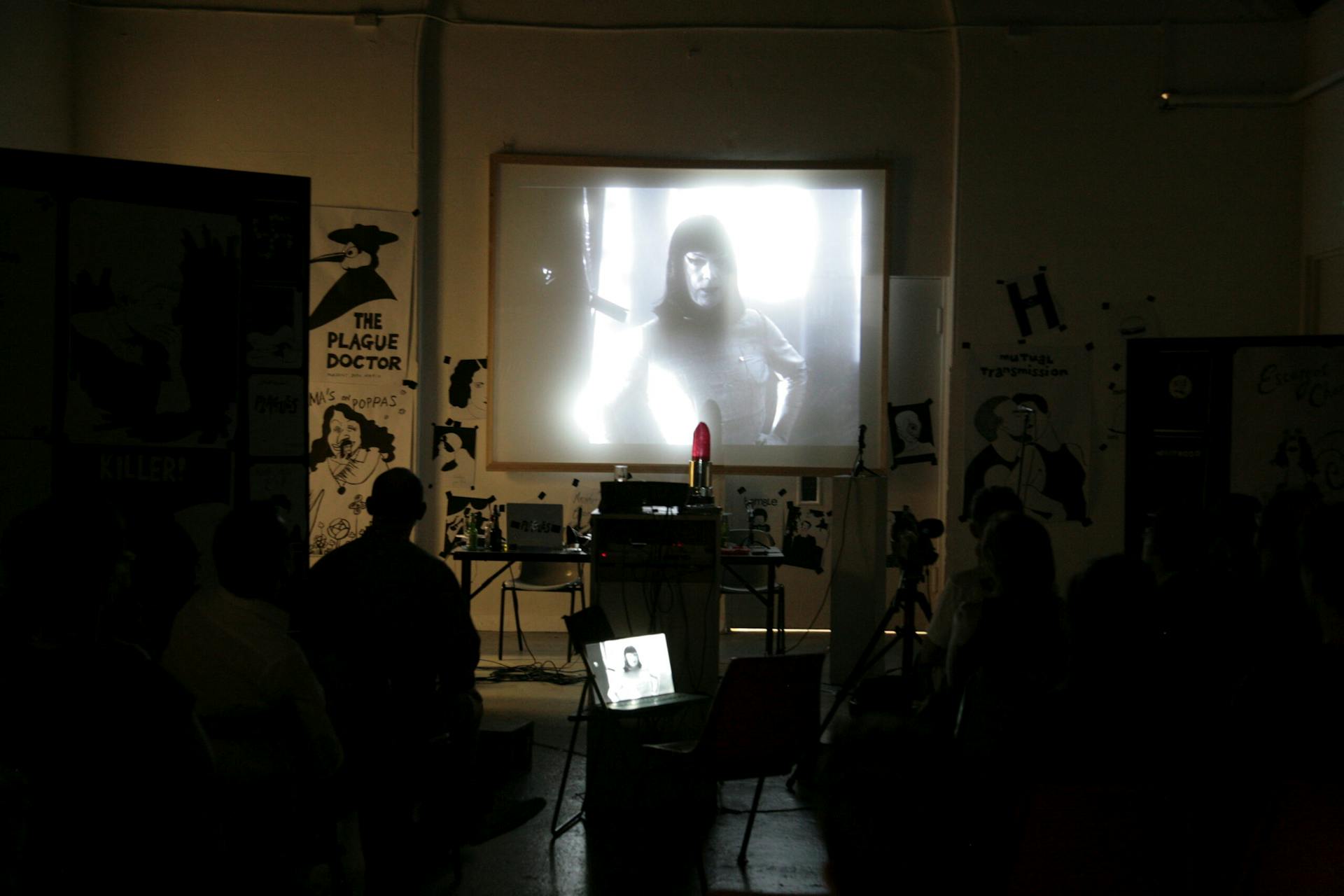 a projection of a black and white film, showing a perfomers in a black wig staring to camera. The film is being screend in front of an audience in a darkened room.