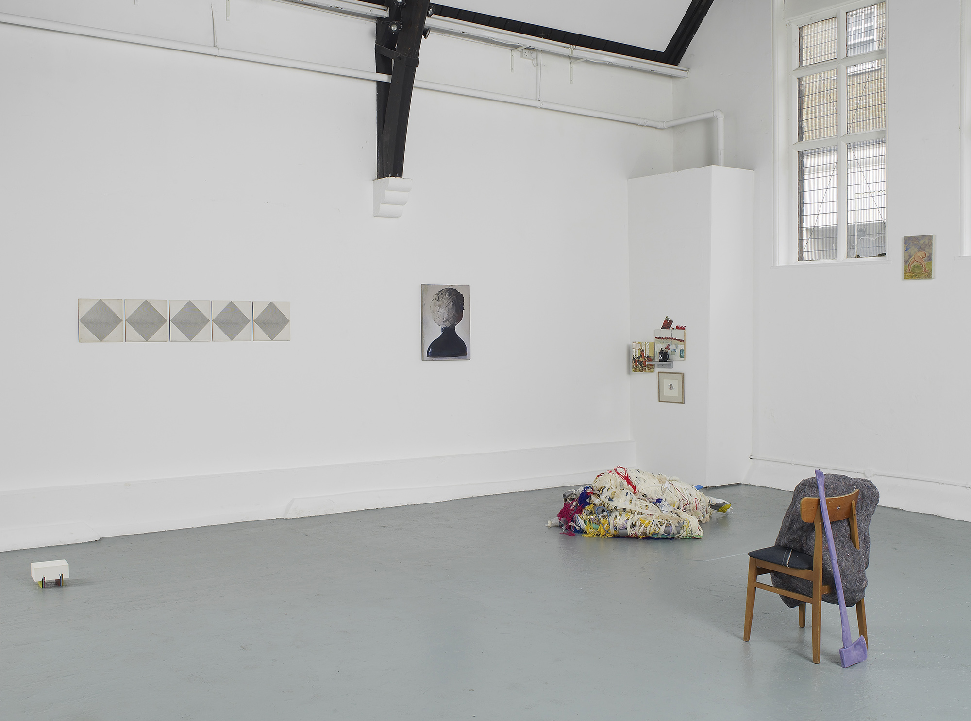 A contemporary art gallery with white walls and high, vaulted ceilings displays sculptures, paintings, and photos. Artworks are placed on the floor, plinths, and walls, in a minimalist, spacious arrangement.