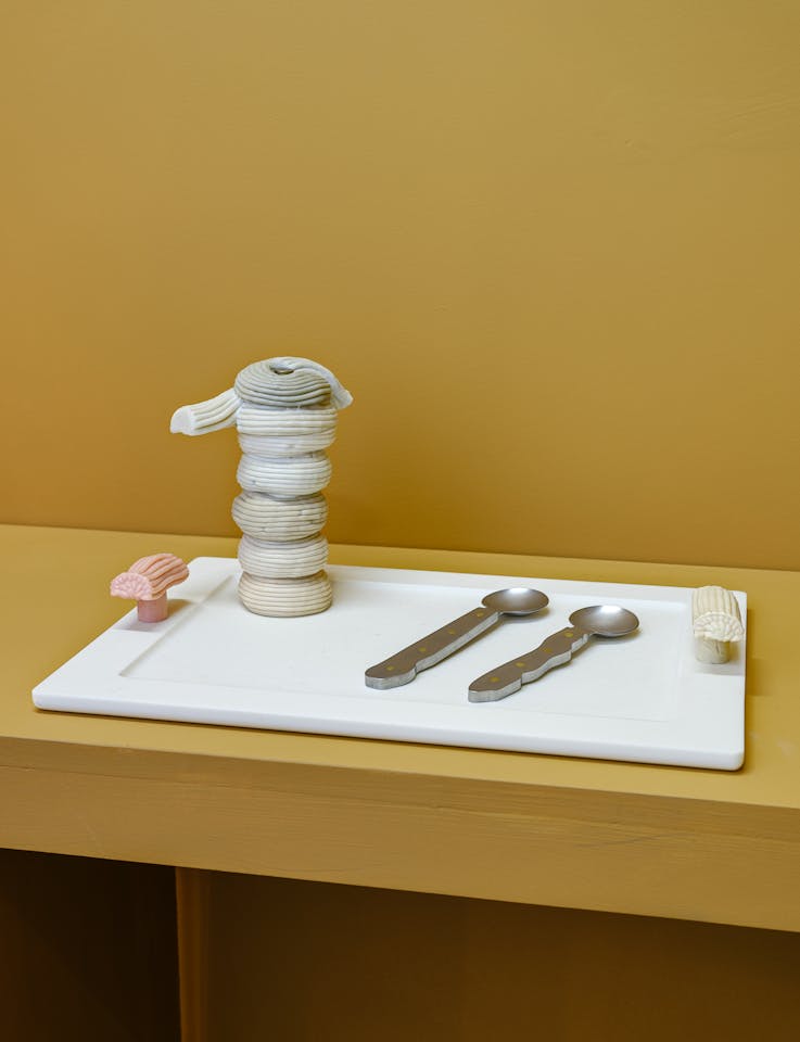 A white tray holds a stack of round, cream-coloured objects created by James Shaw, two metal utensils, and two small, textured items resembling pastries, all set against a mustard yellow background.