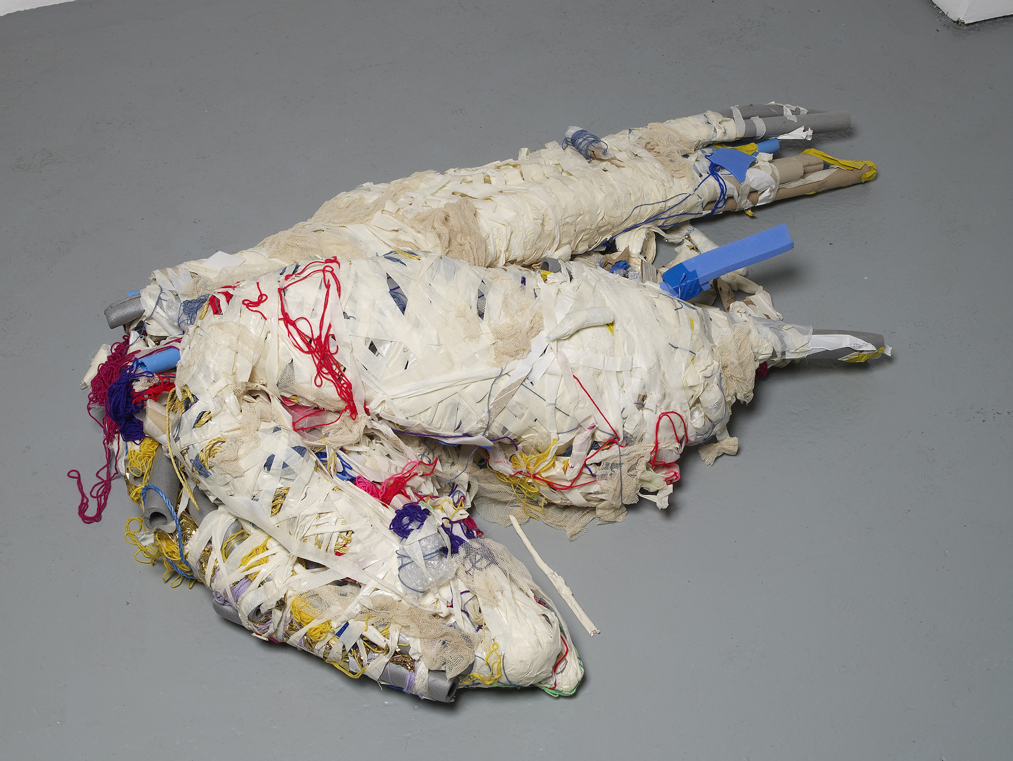 Two large, abstract forms made of white, wrapped fabric and plastic, with red, yellow, and blue threads and tubes, are lying intertwined on a grey floor in an art gallery setting.