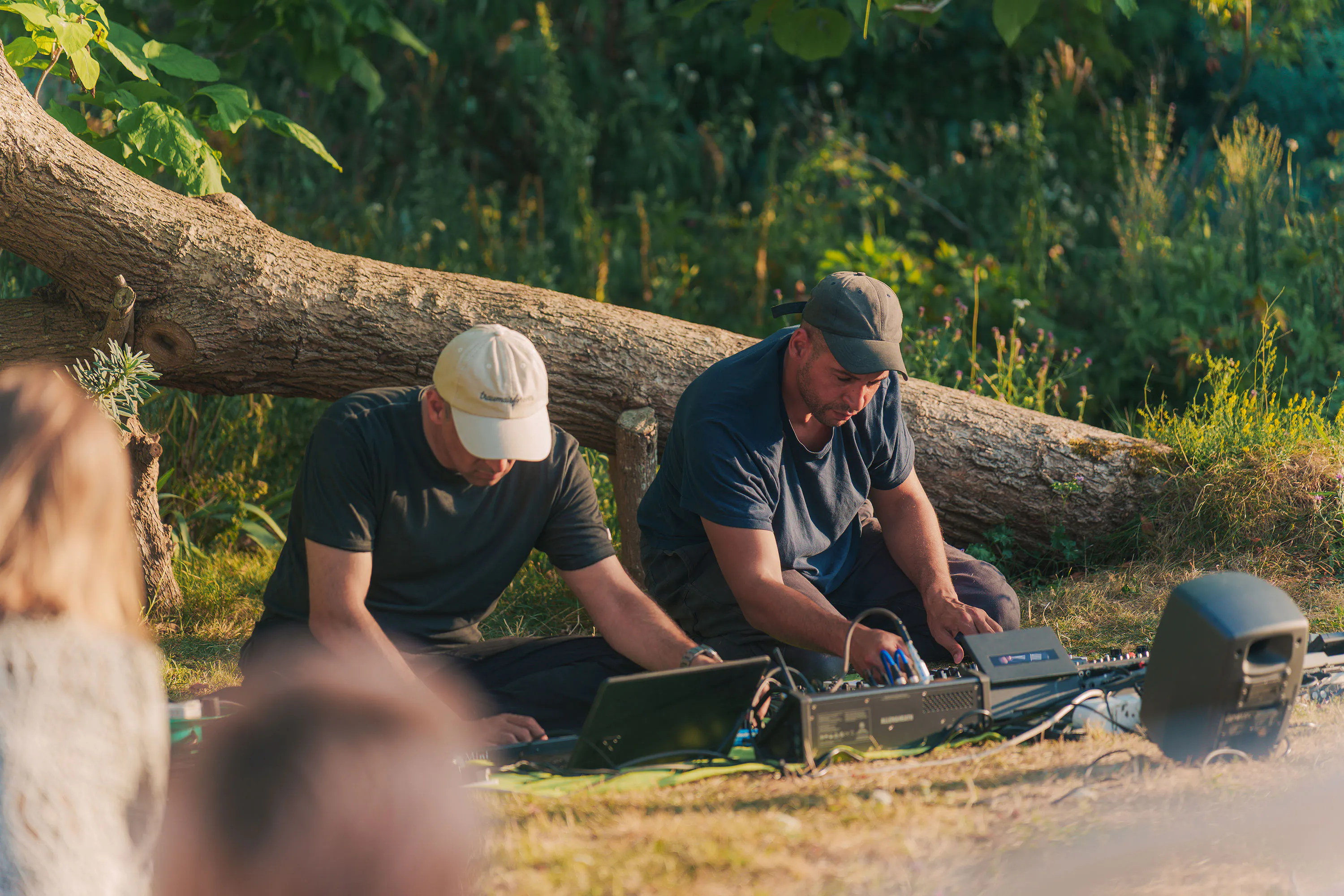 Abbas Zahedi and Tom Harris sit on the grass in front of a fallen log, adjusting audio equipment and a laptop. They are surrounded by foliage, with parts of an audience visible in the foreground.