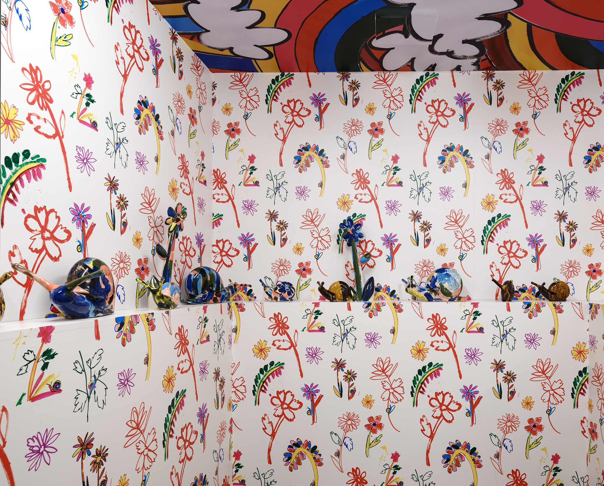 Colourful room with walls and ceiling covered in bright, hand-drawn flower and leaf patterns. A shelf displays abstract and colourful glazed ceramic snails, blending into the vibrant floral wallpaper. The ceiling features rainbow and cloud designs.