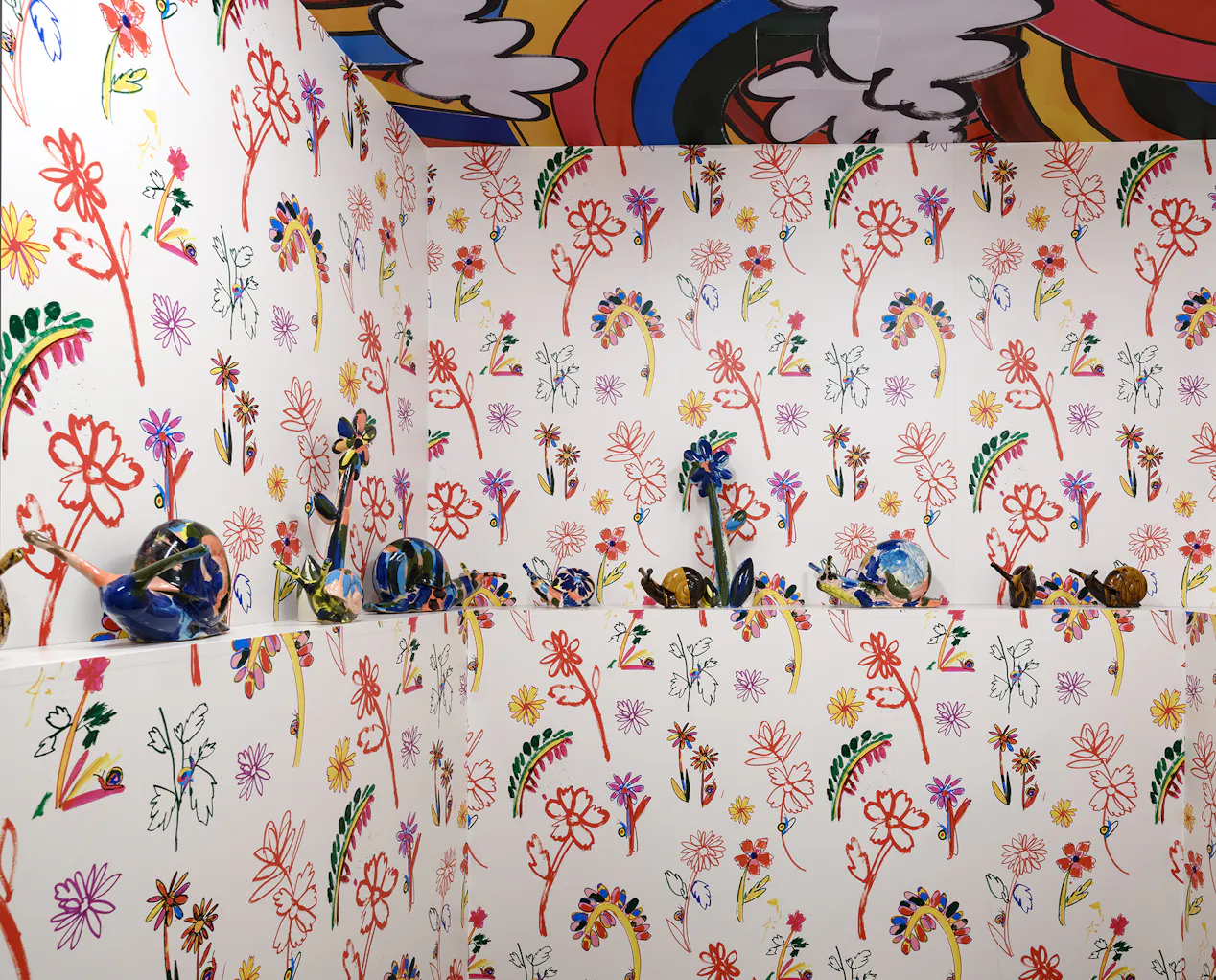 Colourful room with walls and ceiling covered in bright, hand-drawn flower and leaf patterns. A shelf displays abstract and colourful glazed ceramic snails, blending into the vibrant floral wallpaper. The ceiling features rainbow and cloud designs.