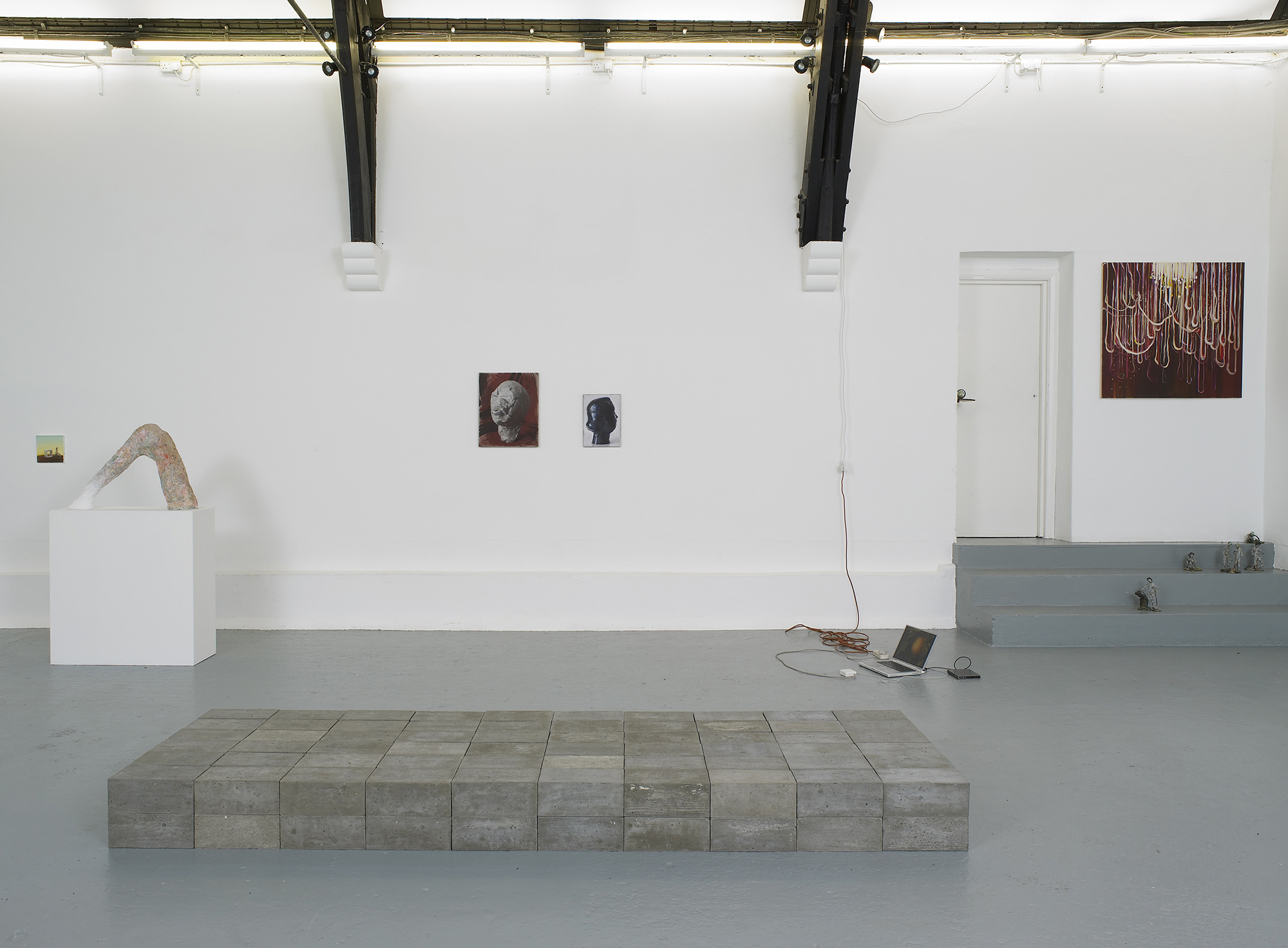 A contemporary art gallery with white walls and high, vaulted ceilings displays sculptures, paintings, and photos. Artworks are placed on the floor, plinths, and walls, in a minimalist, spacious arrangement.