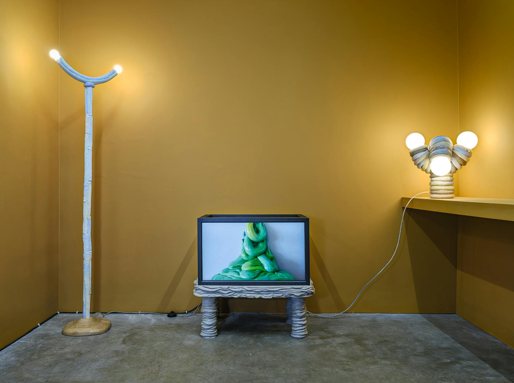 A room with mustard yellow walls features a unique white floor lamp with two glowing bulbs, a TV showing a green gloopy mass on a sculptural white stand, and a matching white table lamp with three bulbs on a ledge.