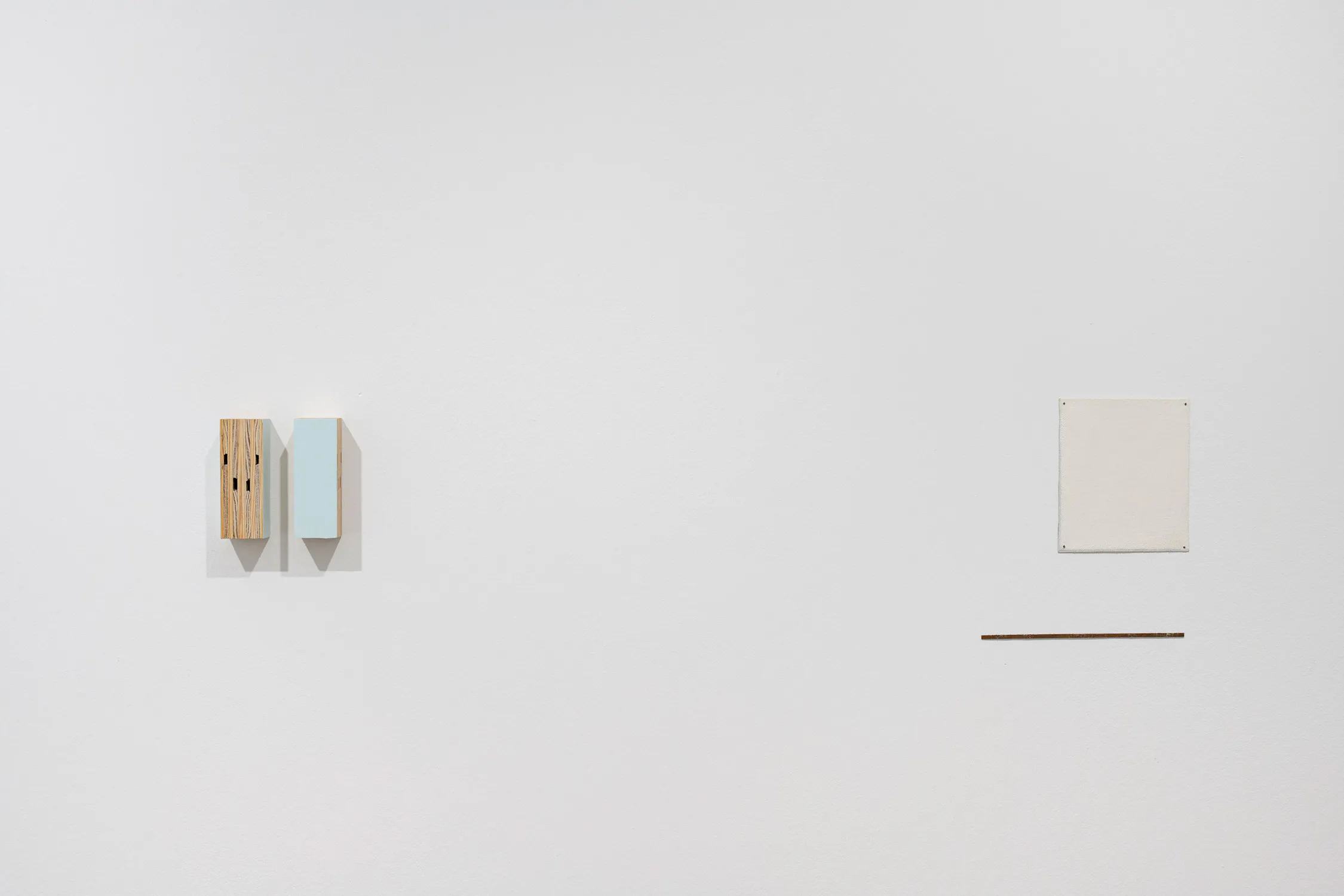 Two small, vertical rectangular blocks—one wood-grained and one light blue—are placed side by side on a white wall. To their right, a square, off-white canvas hangs above a thin, horizontal brown rod.