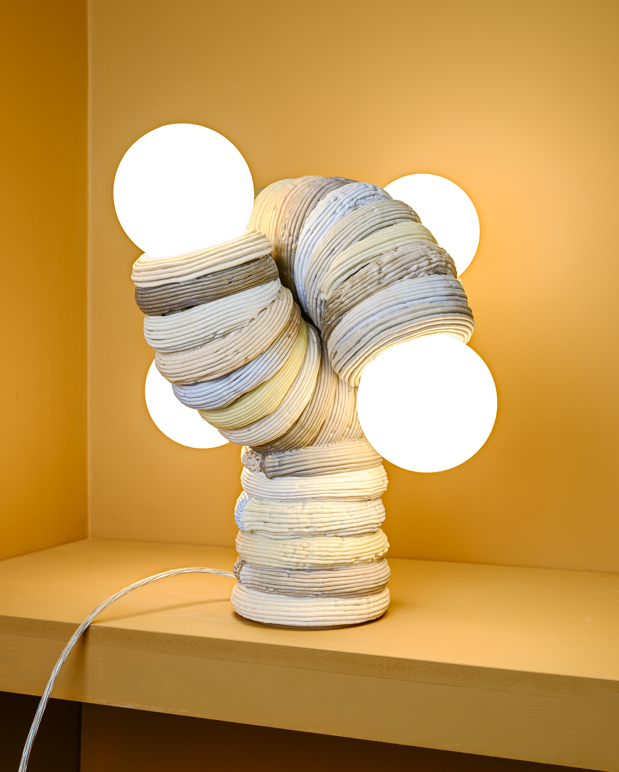 A sculptural table lamp with a curved created by James Shaw, with a ribbed plastic body in neutral tones, featuring four round white bulbs extending outward, is displayed on a yellow shelf against a matching yellow background.
