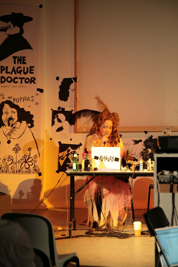 a performer with a red wig and a frilly gown reads from a laptop. The wall in the background is plastered with black and white illustrated posters