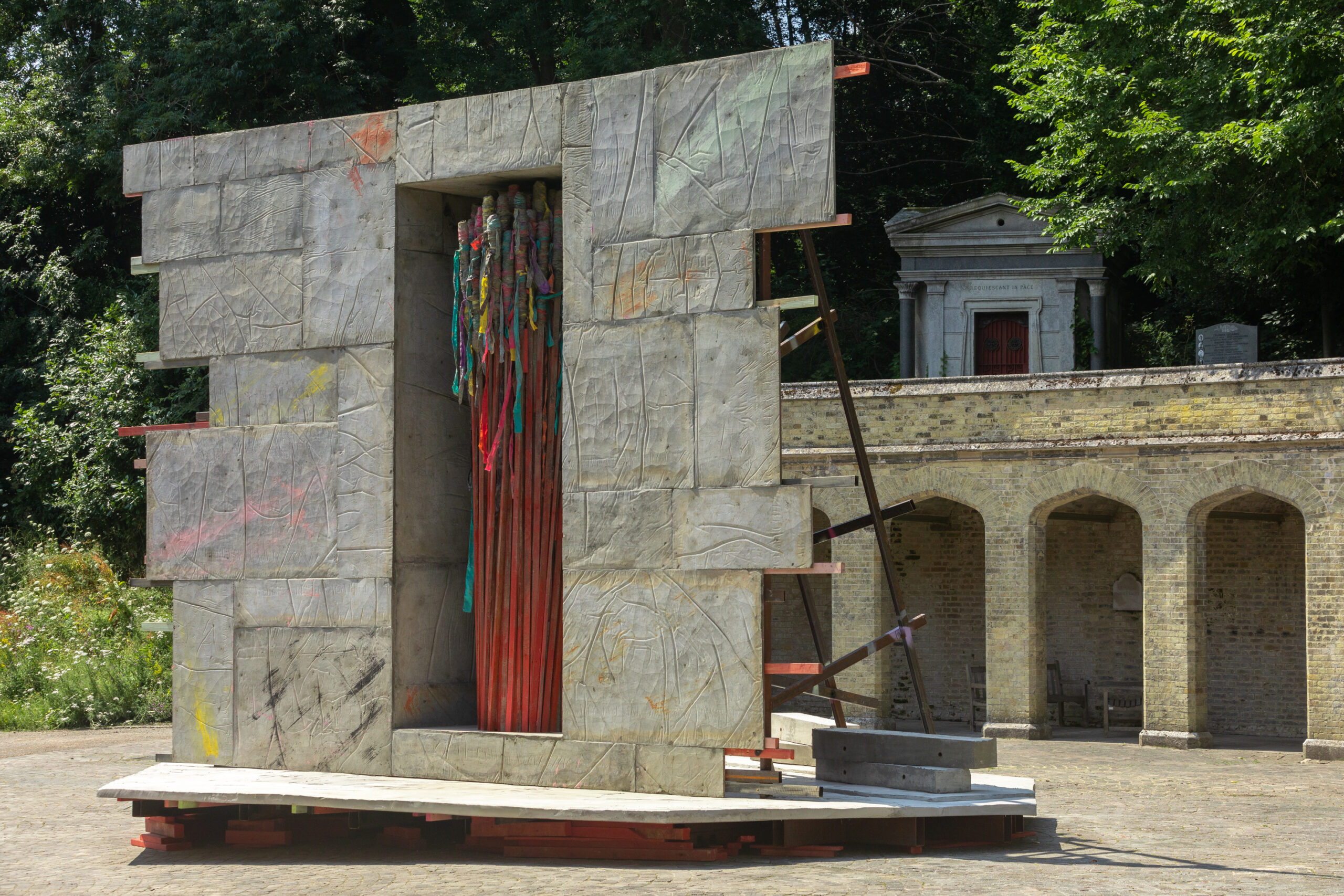 A large, abstract outdoor sculpture made of grey concrete slabs with rough, carved lines stands upright. Its centre is open, revealing red vertical rods with colourful ribbons hanging. Behind it are stone arches and a small building surrounded by greenery.