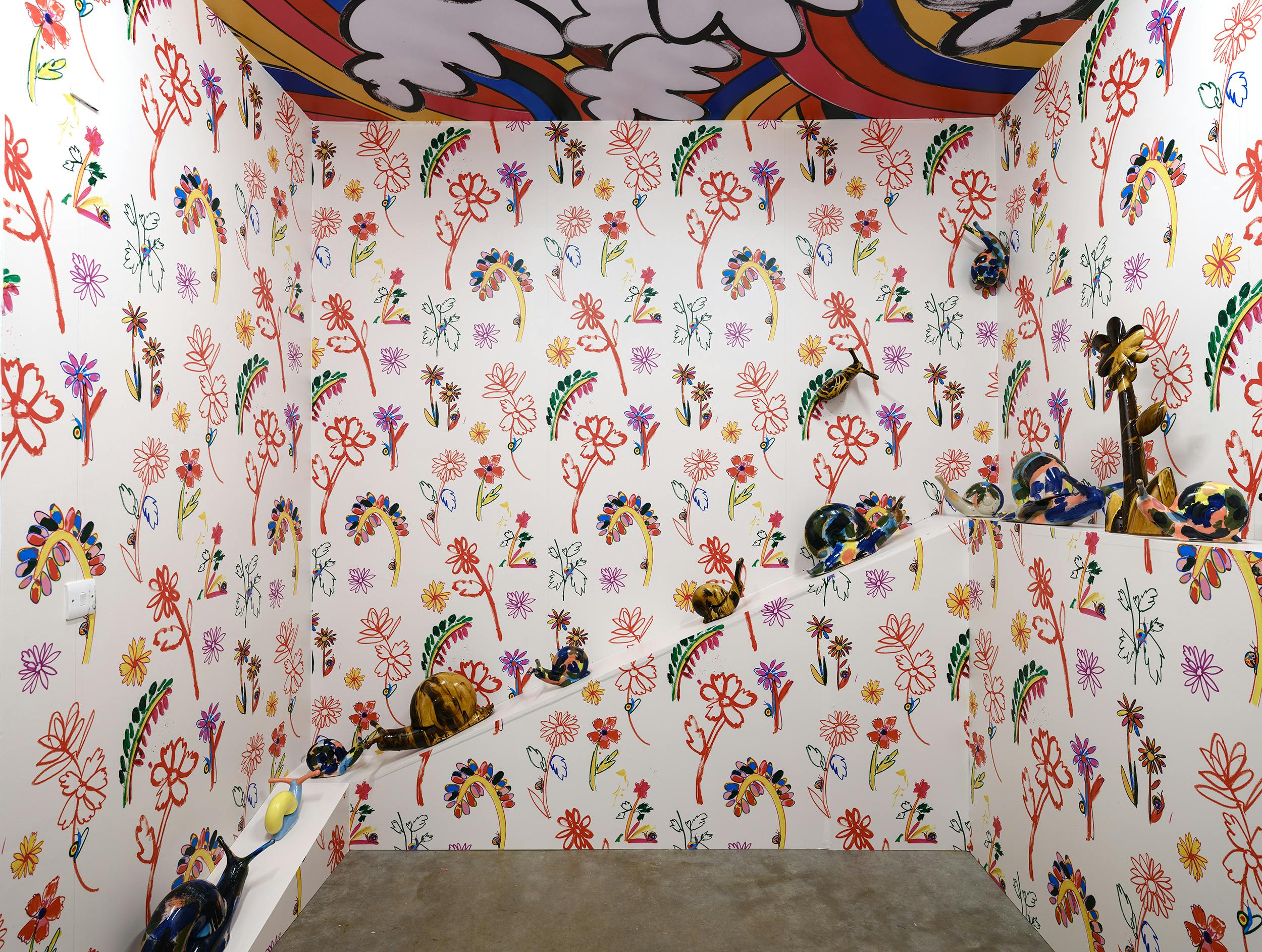 An installation of colourful glazed ceramic snails by Supergroup set against a white wall covered in vibrant, hand-drawn floral and abstract patterns. The floor is concrete, and the ceiling edge also features colourful designs.