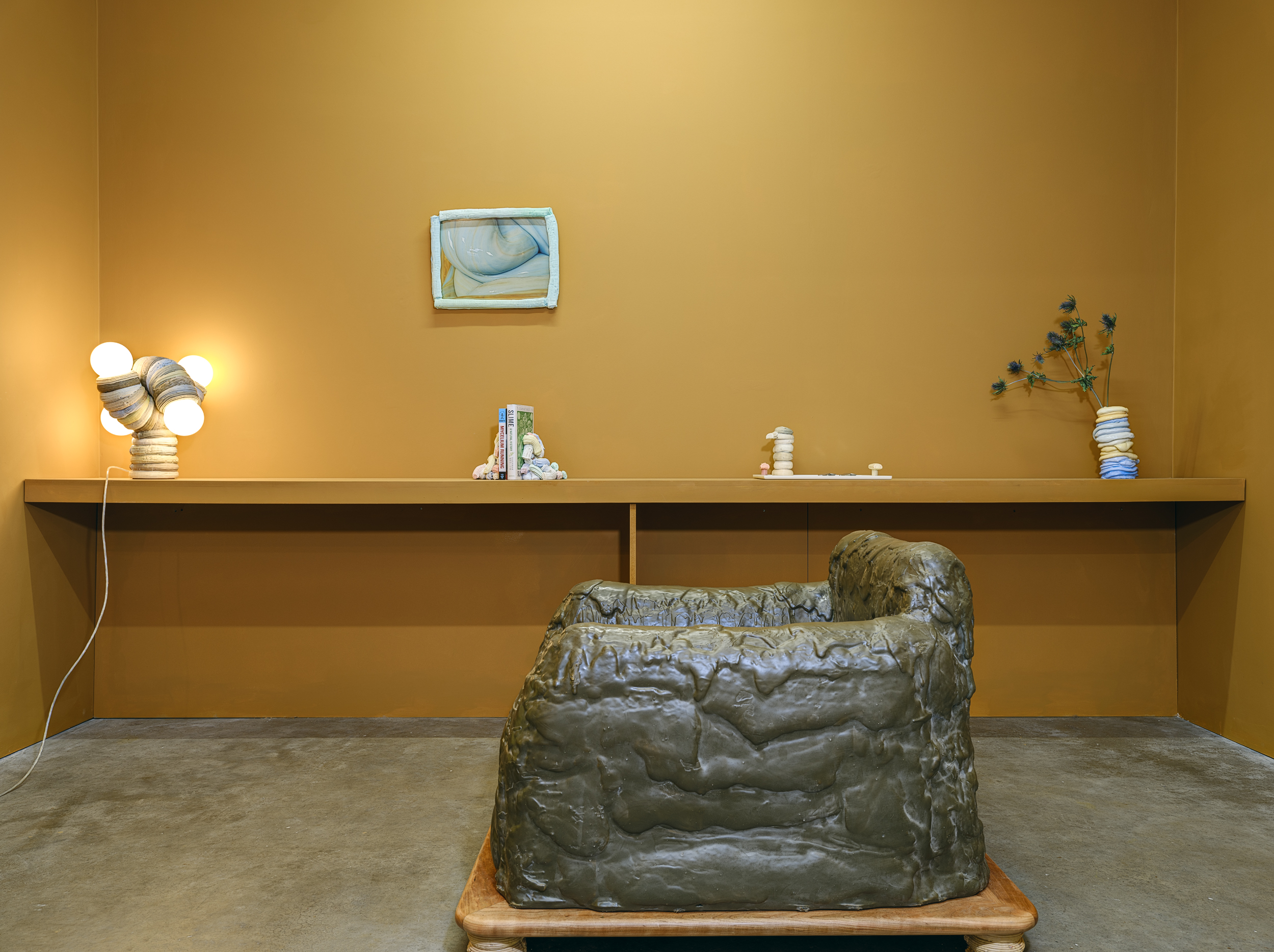 A modern room with mustard yellow walls, a sculpted, stone-coloured plastic armchair, a long shelf with a lamp, a small painting, books, decorative objects, and a vase with flowers, all on a concrete floor.